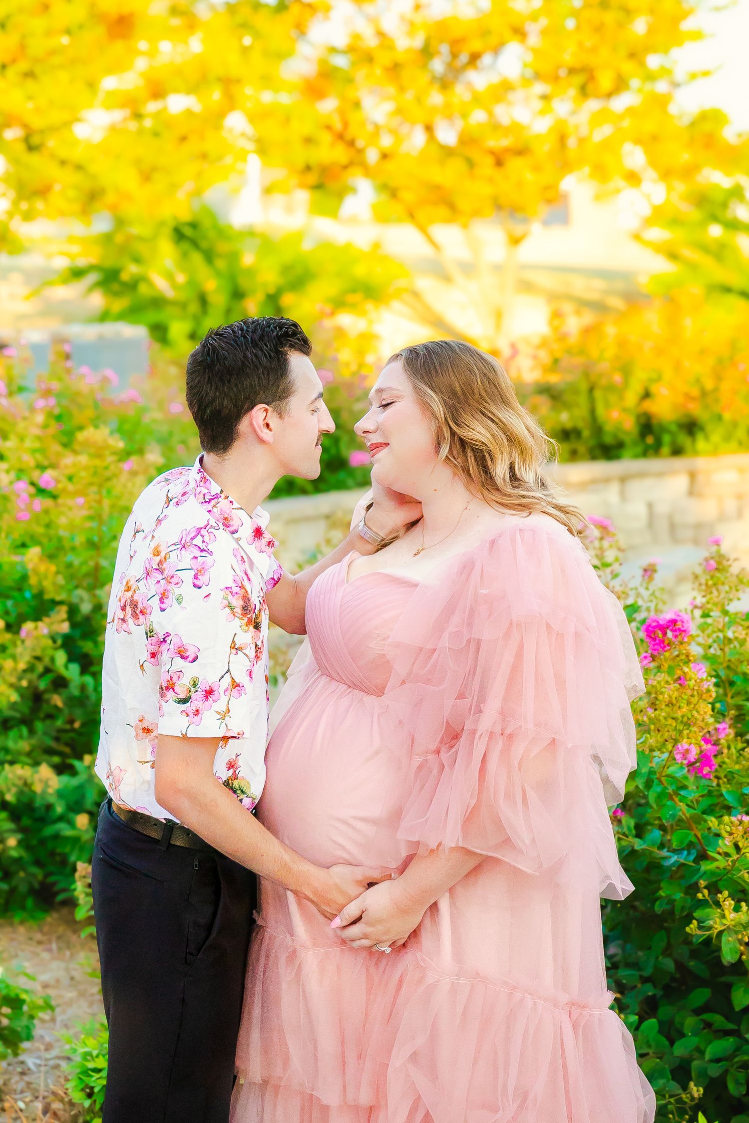 A romantic maternity photo taken in autumn with vibrant yellow foliage and pink flowers in the background.