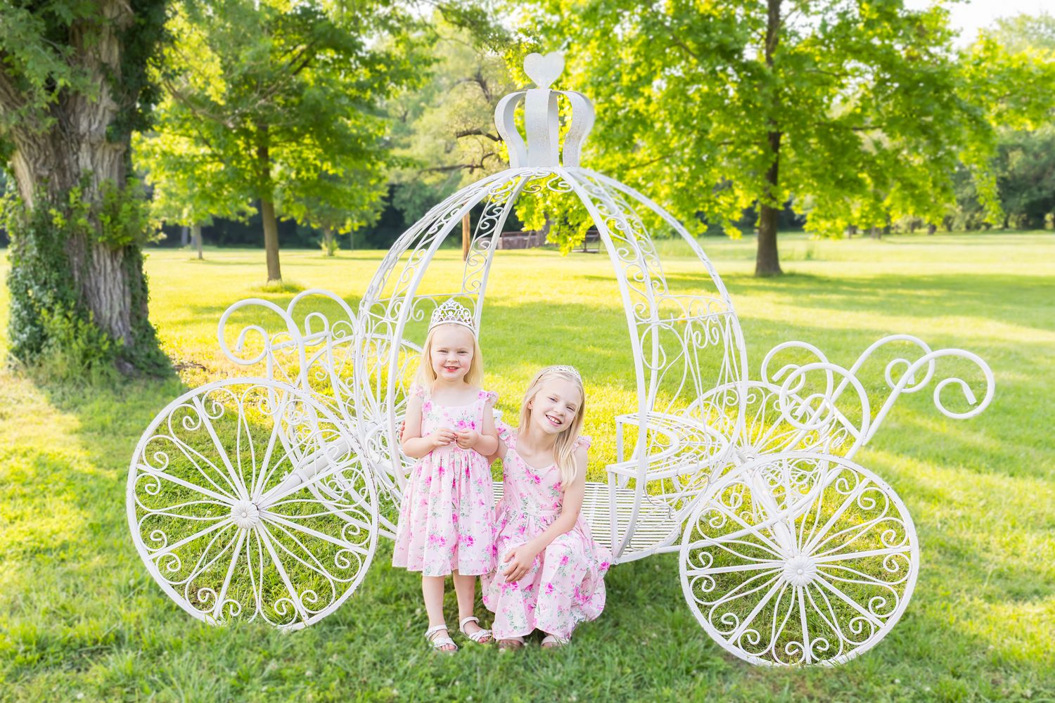 Two little girls with matching pink dresses smile in front of a large white carriage during a mini session photoshoot in Pittsburg Kansas.