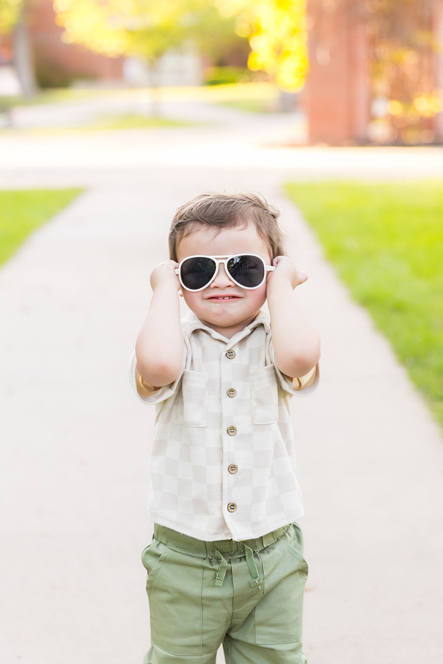Child wearing sunglasses, white button-up and green pants outdoors for two year old photoshoot at Pitt State University in Pittsburg KS.