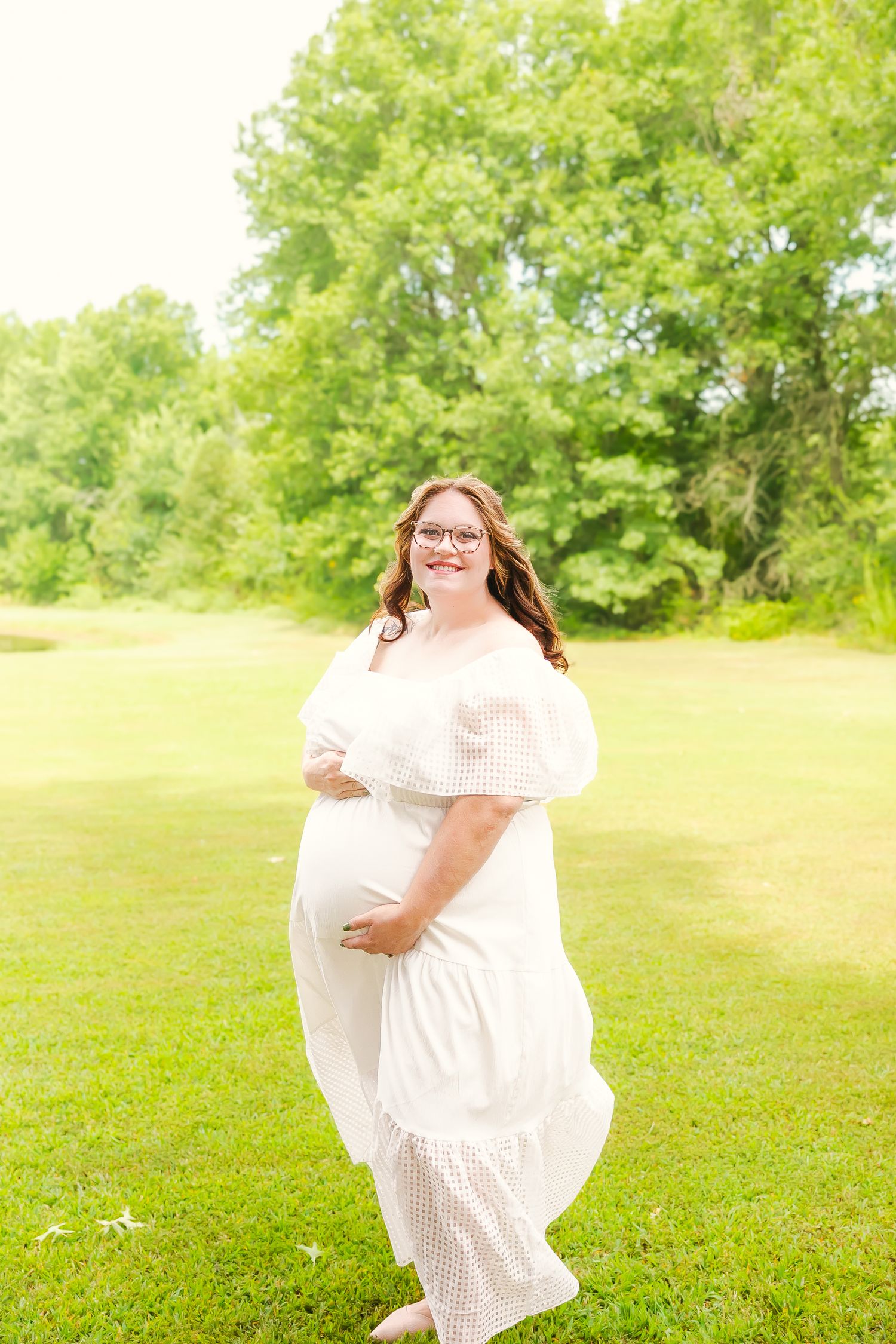 A person in a flowing white dress poses outdoors on green grass with trees in the background.