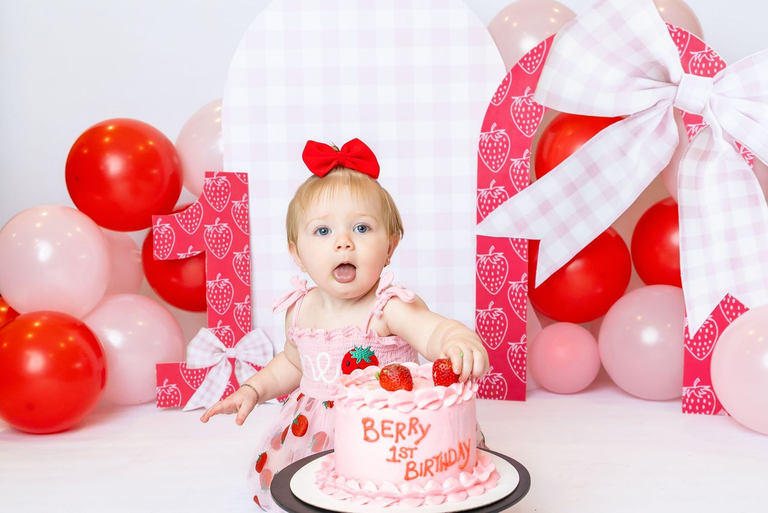 Baby sits in from of a cake grabbing strawberry off it with her tongue out for her 1st birthday photoshoot with Annie Robinson Photography.