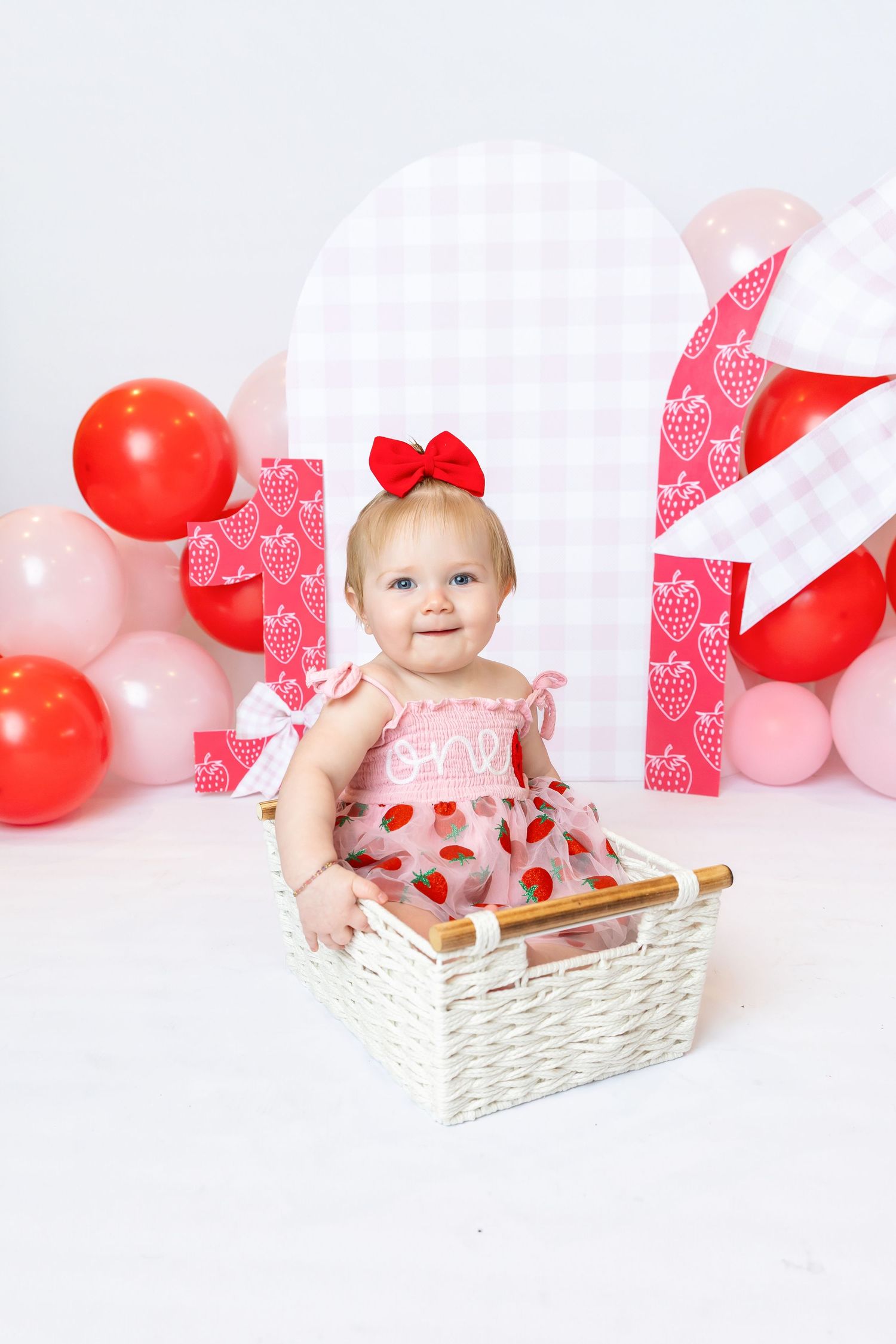 Baby grins in basket wearing a strawberry pink dress during 1st birthday shoot with Annie Robinson Photography of Pittsburg KS and Joplin Mo