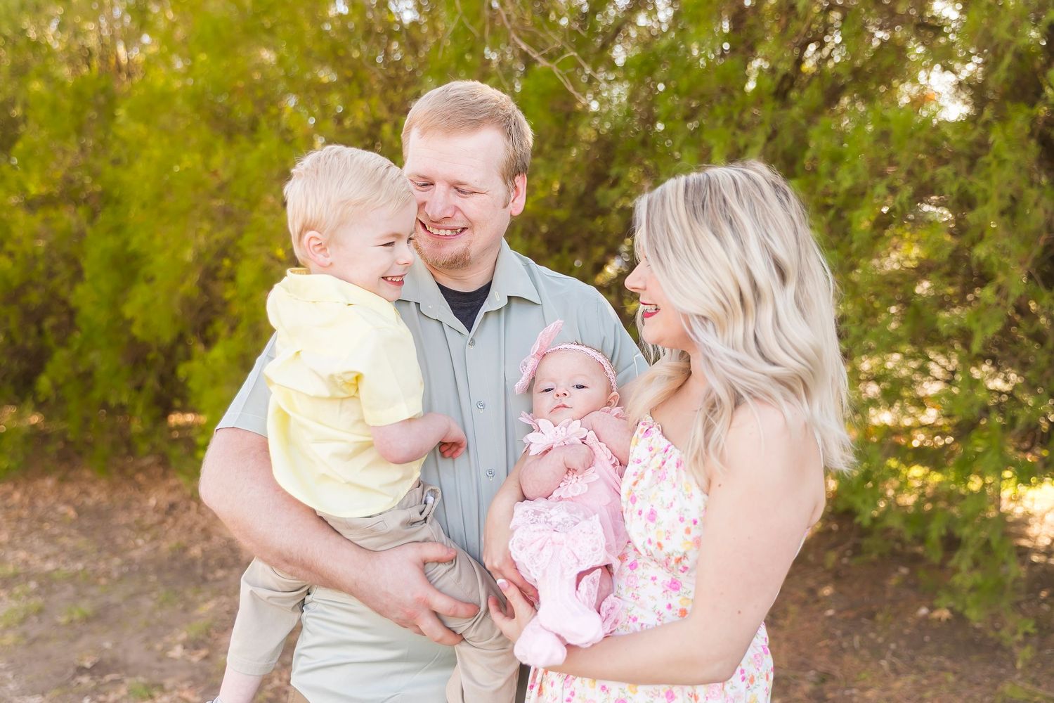 A happy family group wearing pastel colors poses together outdoors against natural greenery in warm sunlight.