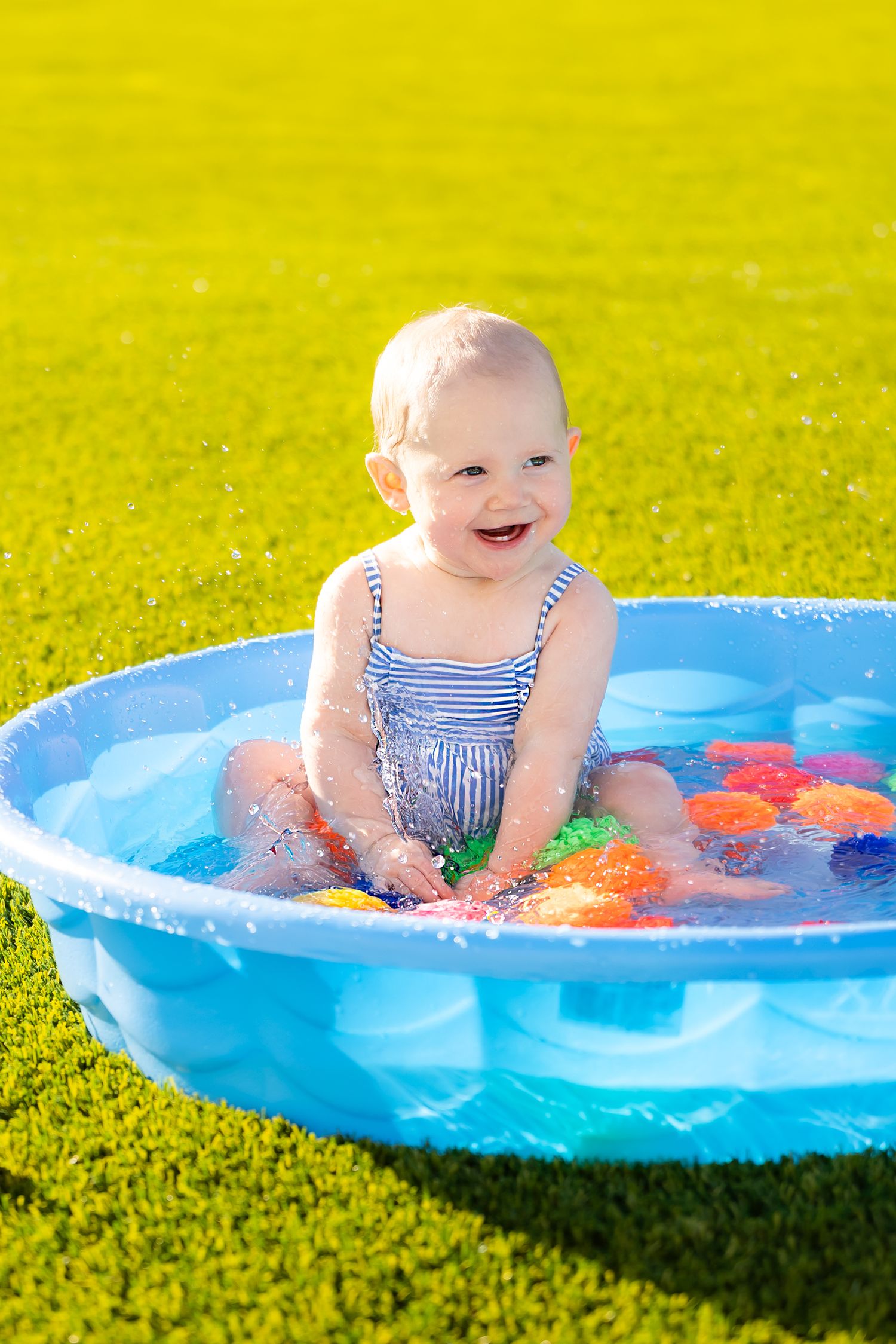 Smiling toddler in blue striped swimwear playing in a kiddie pool with floating toys during summer mini session photoshoot in Pittsburg KS.