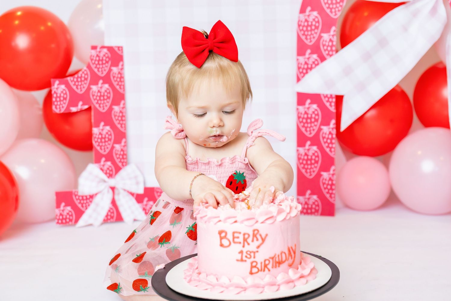 Baby wearing strawberry dress sits in front of cake digging in with her hands for 1st birthday photoshoot with Annie Robinson Photography.