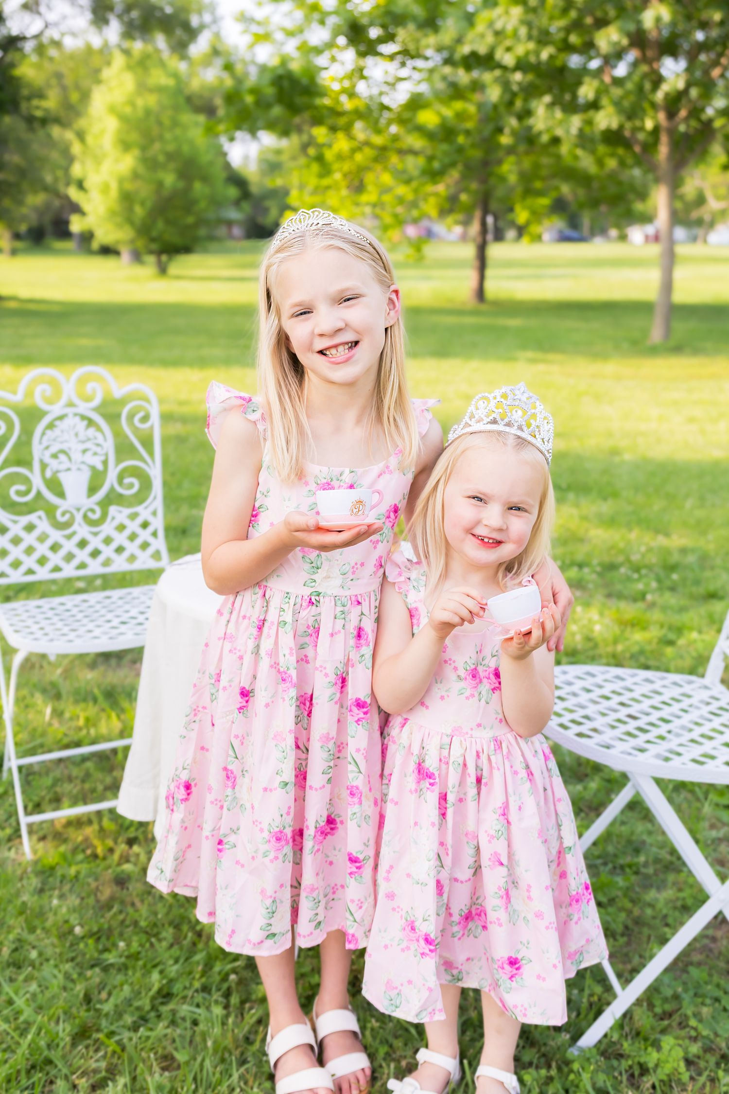 Children in matching pink floral dresses and tiaras smile while standing with tea cups during a mini session photoshoot in Pittsburg Kansas.