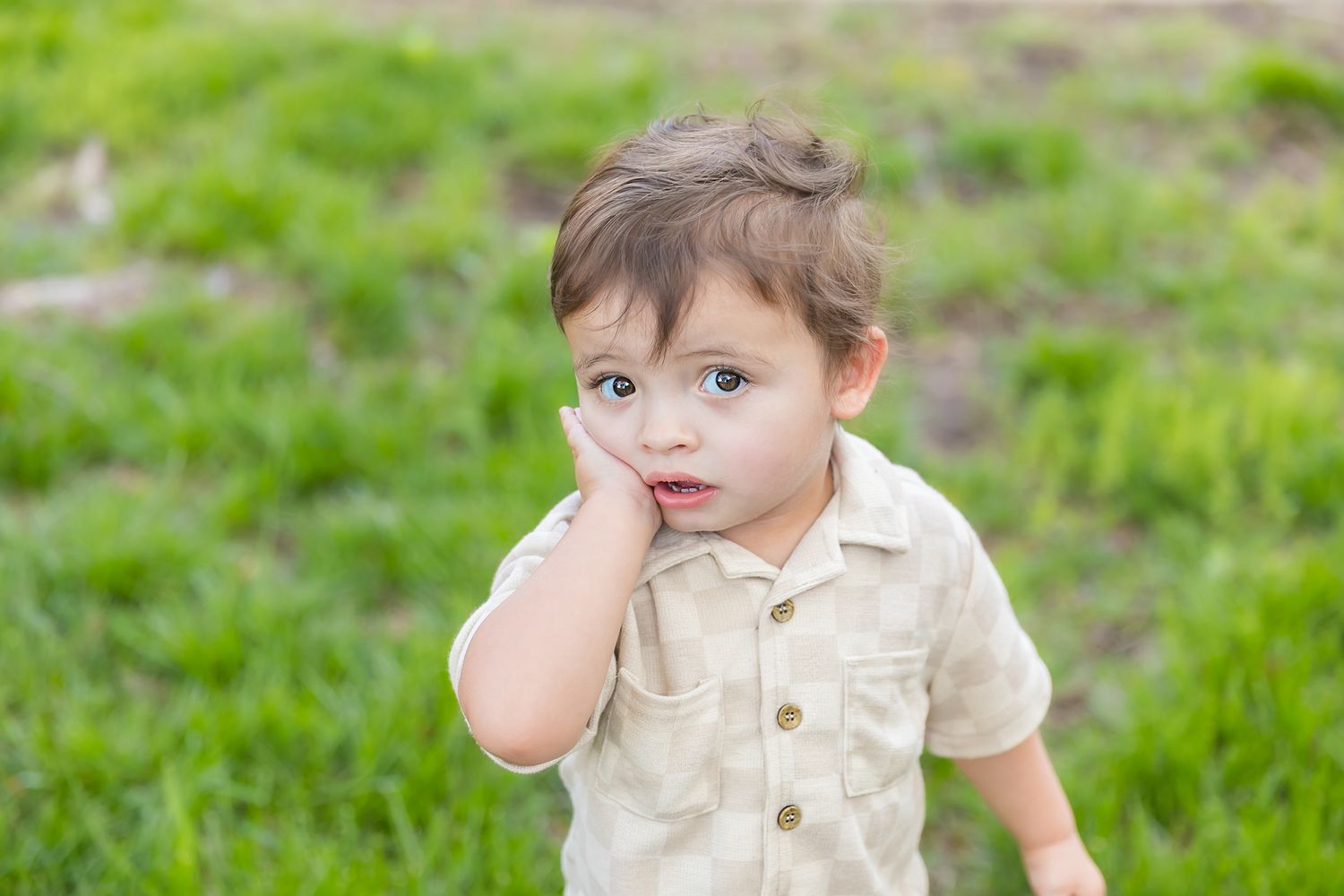 Child in checkered shirt is outdoors in grass with a hand against his cheek creating surprised face for milestone photos in Pittsburg KS.