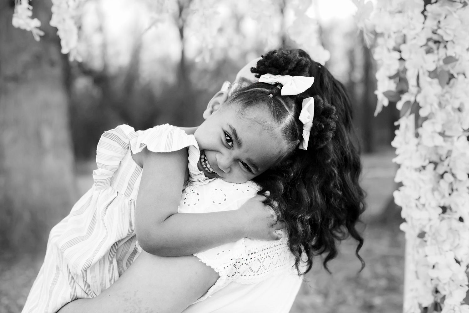 A joyful child in a white dress and bow leans to the side while posing outdoors in a black and white portrait.