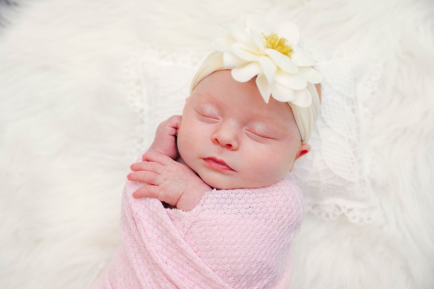Newborn baby swaddled in pink blanket with white daisy headband sleeping peacefully on soft white fur.