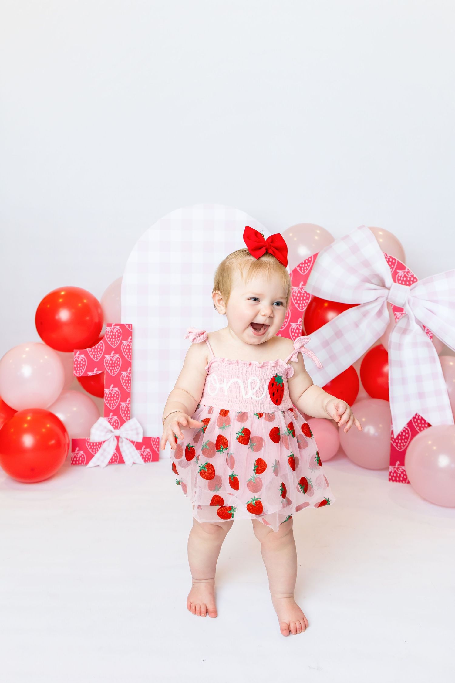 Baby wearing pink strawberry dress stands in front of strawberry setup during 1st birthday photoshoot with Annie Robinson Photography.