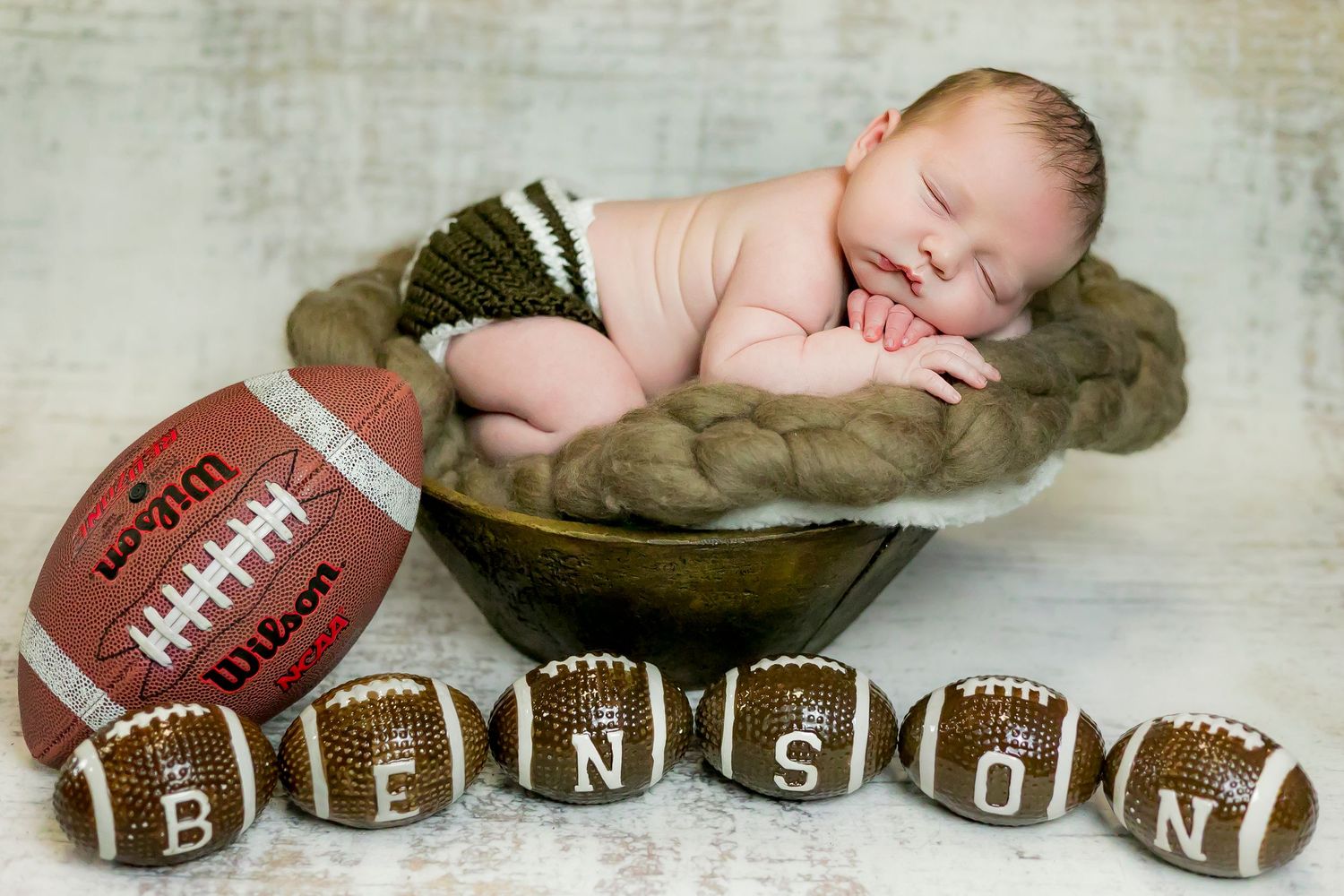 Newborn sleeping on olive green blanket with football and letter blocks spelling out their name below.