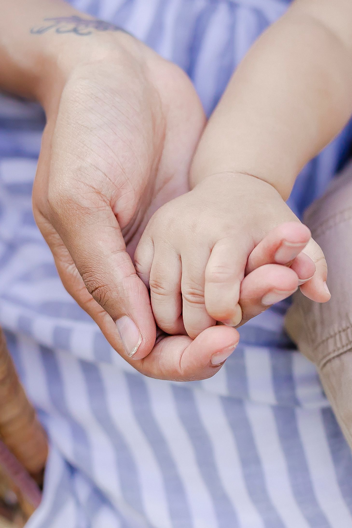 Adult hand gently holds tiny baby hand against light blue striped fabric background.