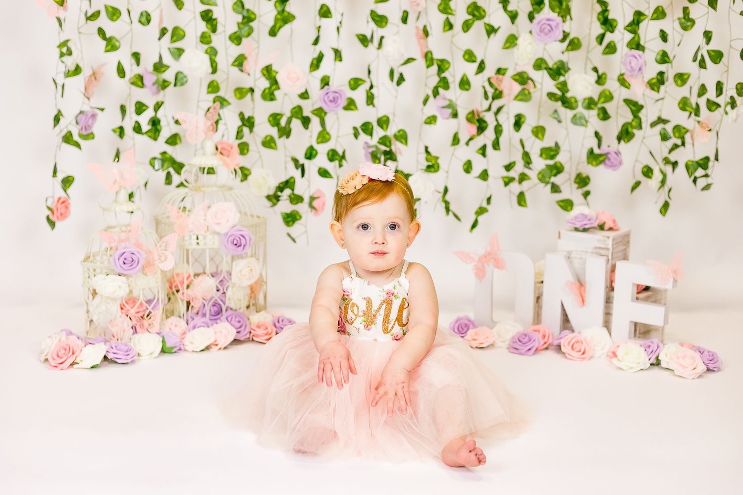 Pretty first birthday setup with floral decorations, leafy vine backdrop, and baby in peach tulle dress sitting on floor.