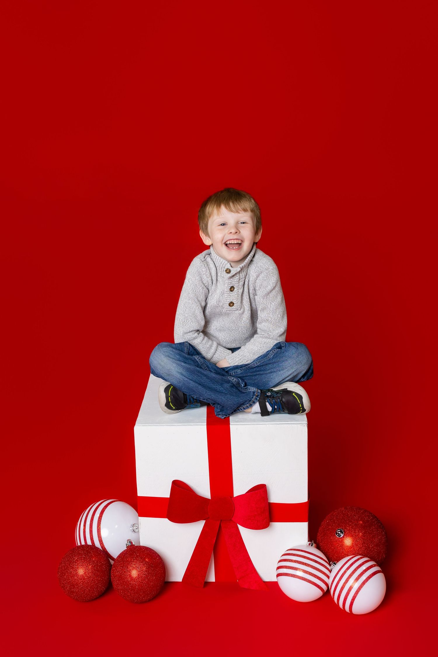 Child sits crisscross on a large white and red gift box, with red and white ornaments for Christmas photos in Pittsburg KS and Joplin MO.