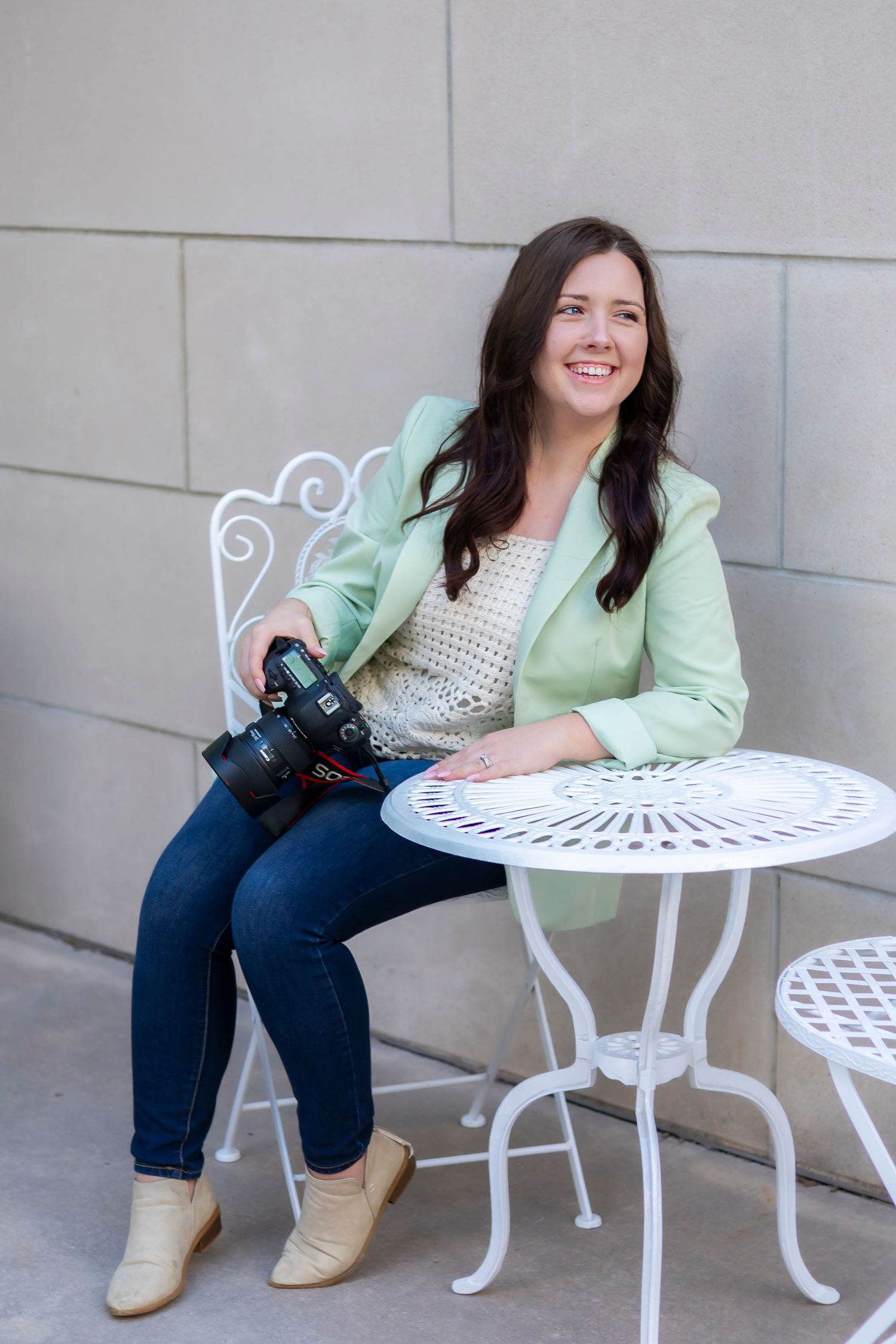 A photographer in Pittsburg KS and Joplin MO with a camera sits at a white wrought iron patio table wearing a mint green blazer and jeans.