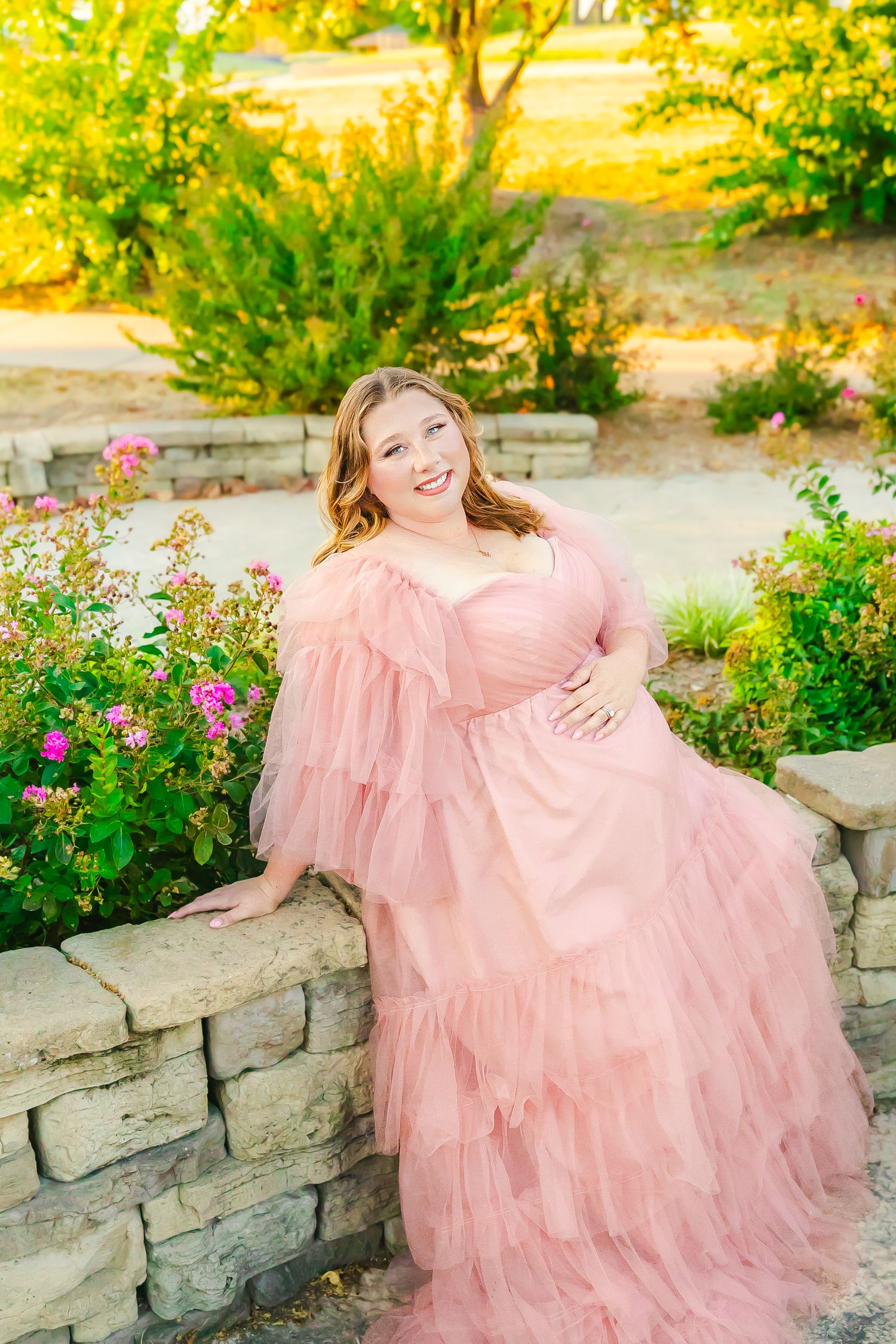 A beautiful pregnant woman in a soft pink tulle gown sits in a garden setting with blooming flowers to pose for a maternity photoshoot.