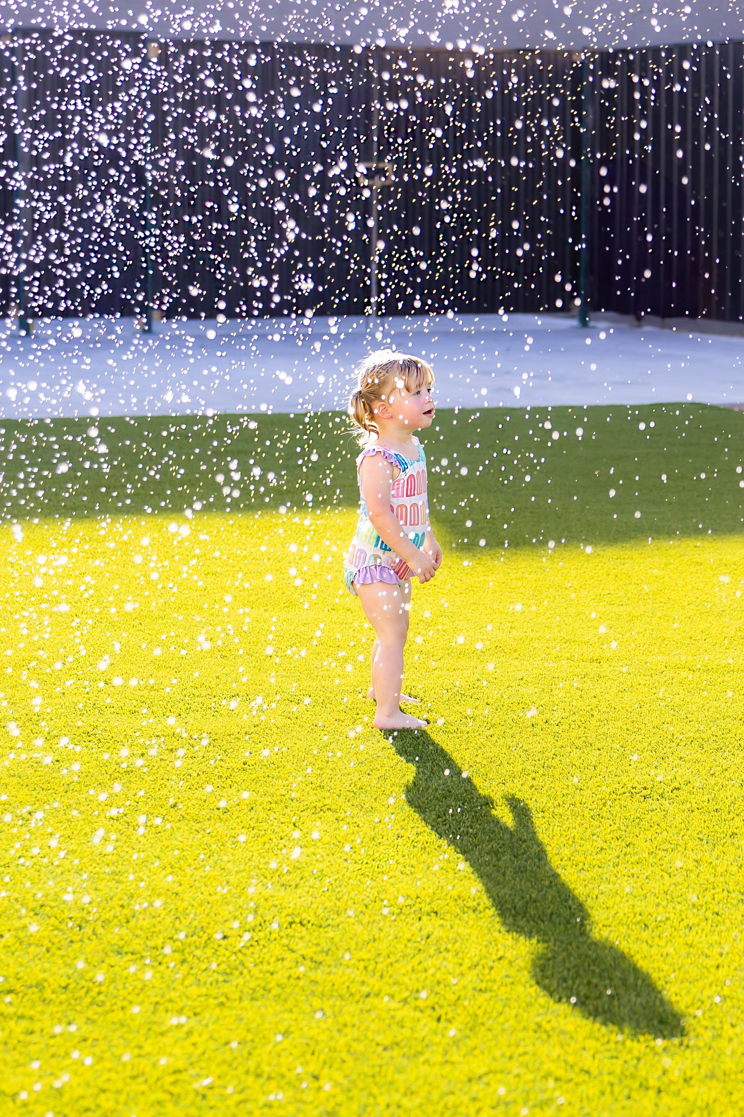 Small child plays joyfully in water sprinklers on a sunny lawn casting a long shadow during a summer mini session photoshoot in Pittsburg KS