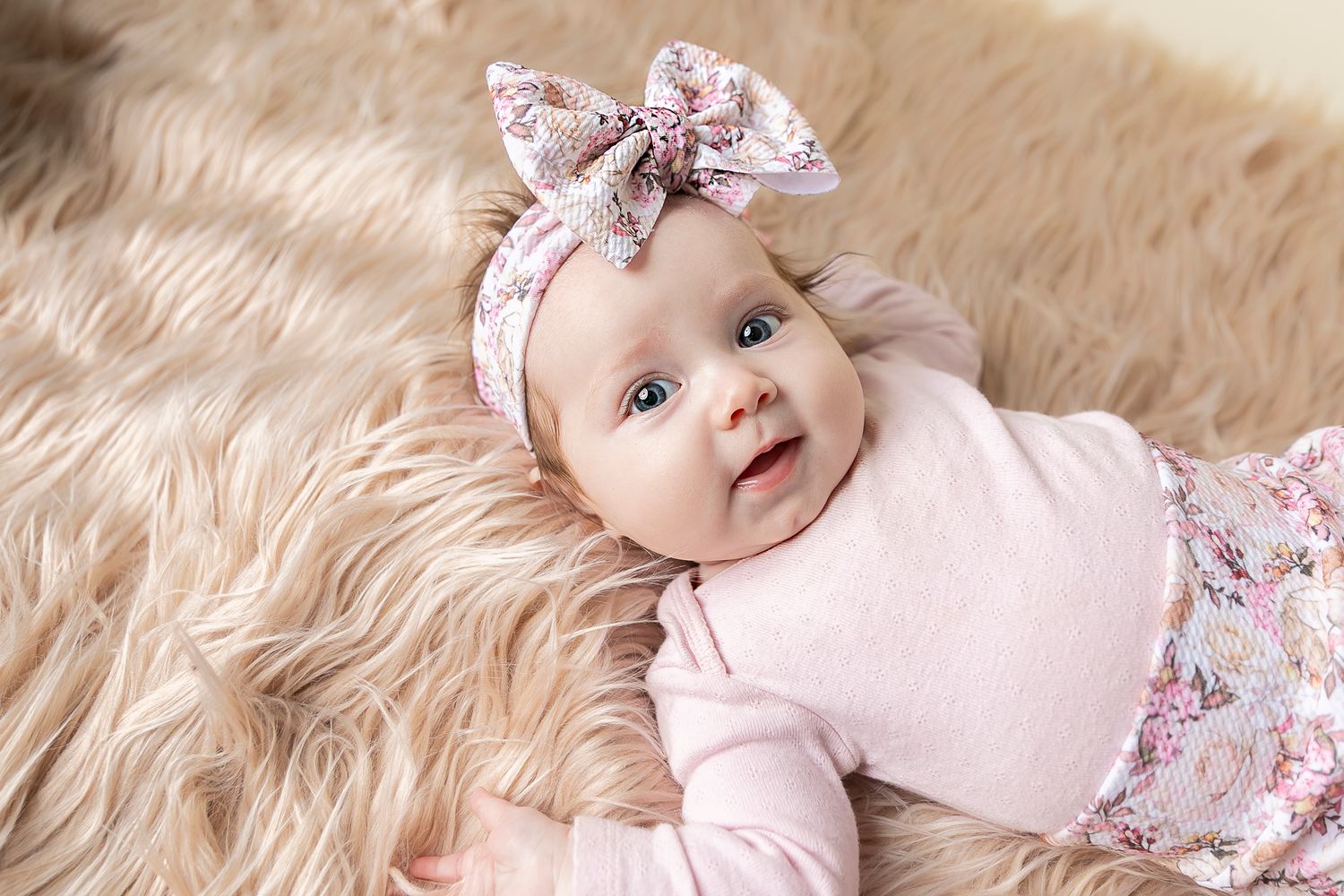 Small infant wearing pink top and floral headband lying on soft fluffy beige fur rug.