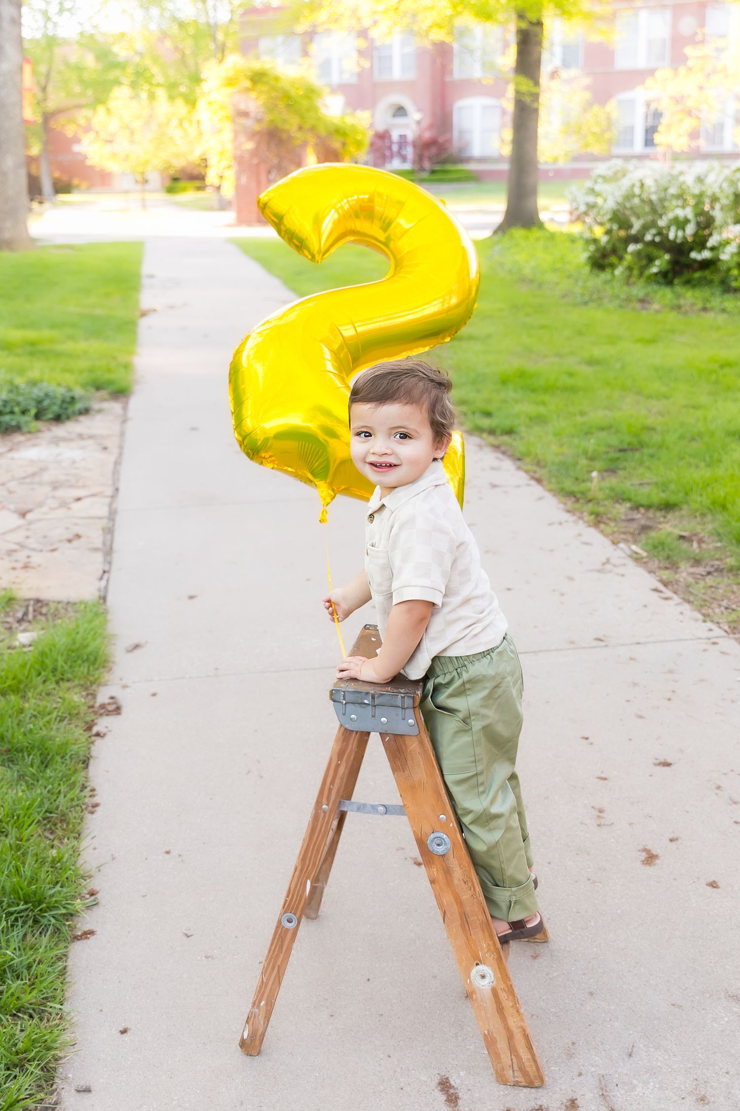 A toddler holds two shaped balloon standing on a small ladder while smiling for birthday photos at Pitt State University in Pittsburg KS.