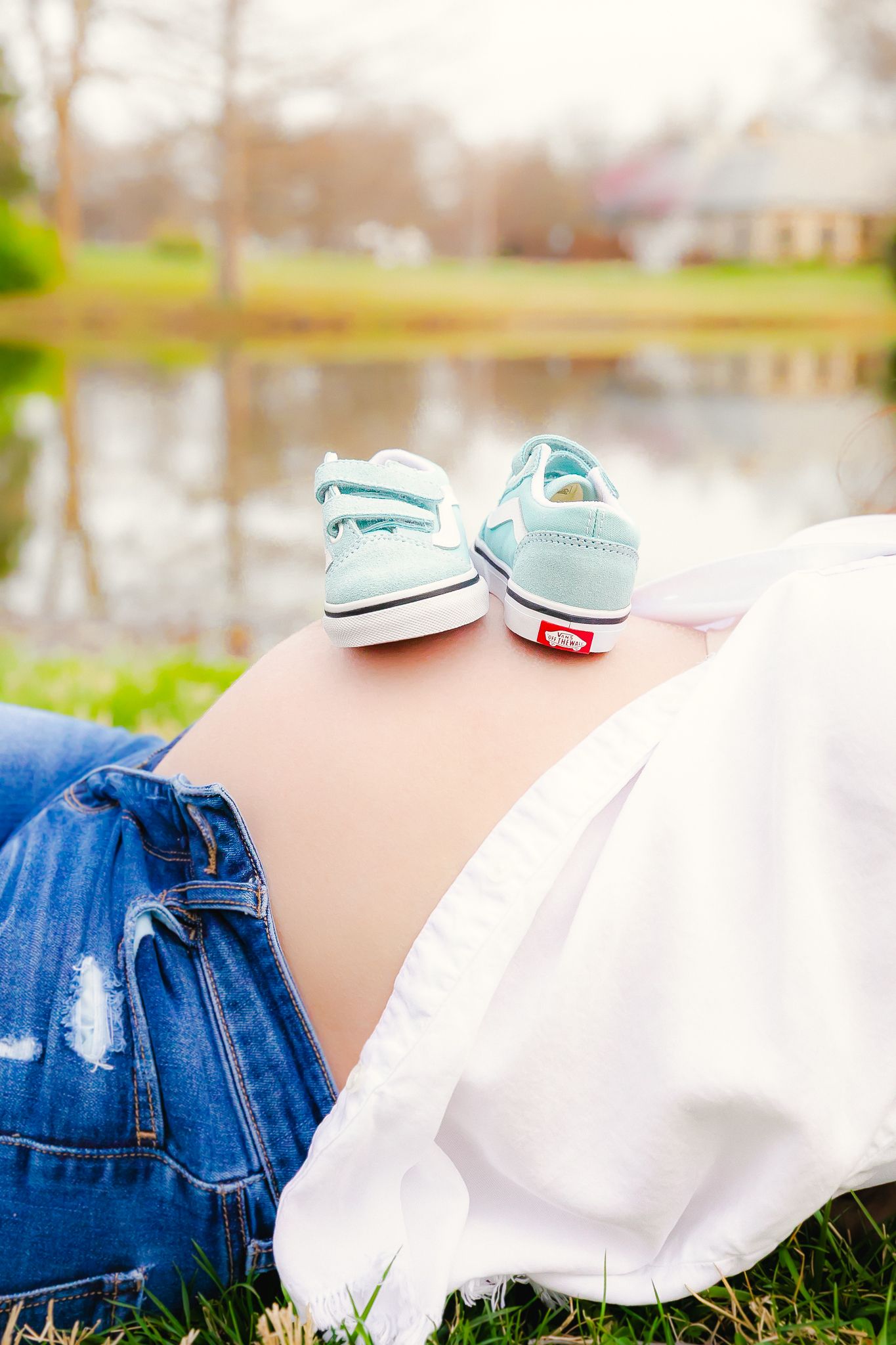 Tiny turquoise baby shoes rest on pregnant belly with white shirt and denim in outdoor setting near pond.