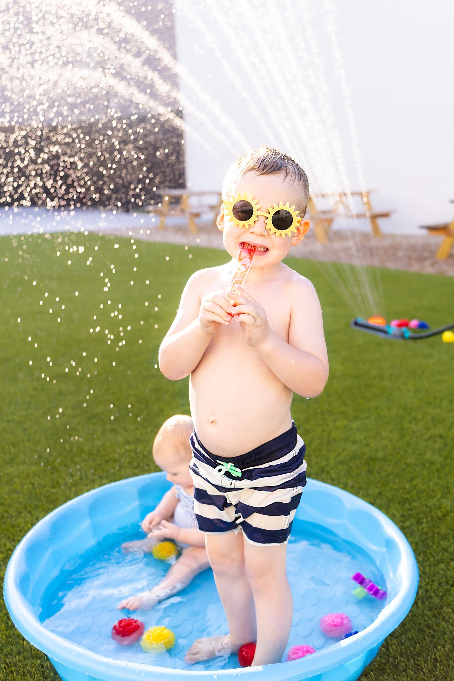 Child wears sunglasses and striped swim trunks enjoying a popsicle in a kiddie pool during summer mini session photoshoot in Pittsburg KS