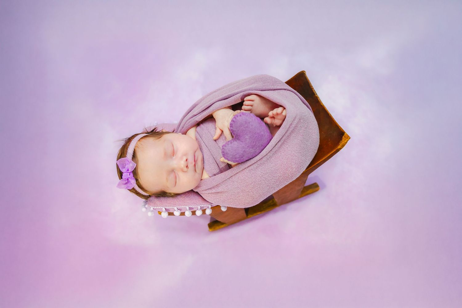 Baby peacefully sleeping in wooden cradle wearing purple outfit against lavender background.
