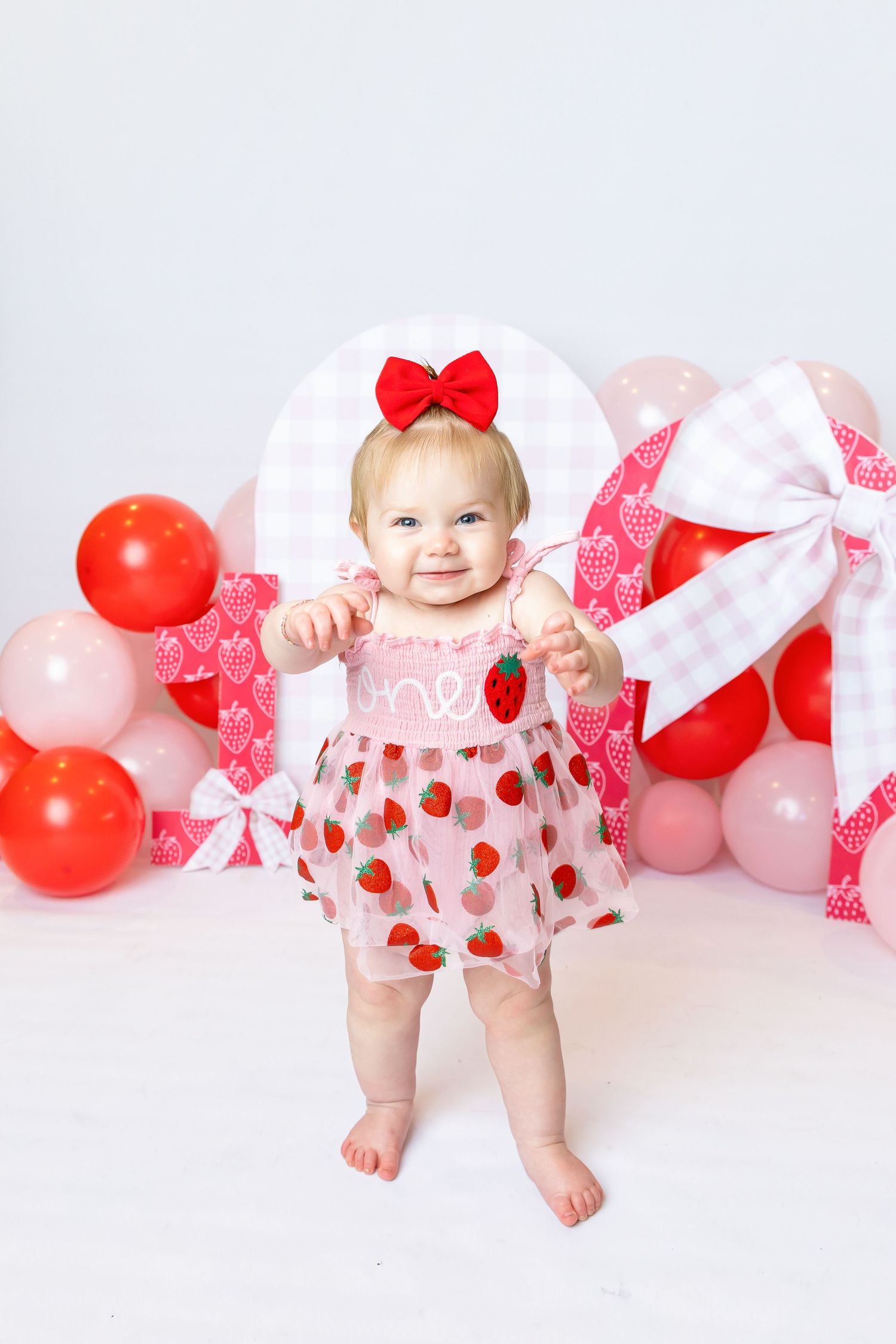 Baby wearing pink strawberry dress stands in front of strawberry setup during 1st birthday photoshoot with Annie Robinson Photography.