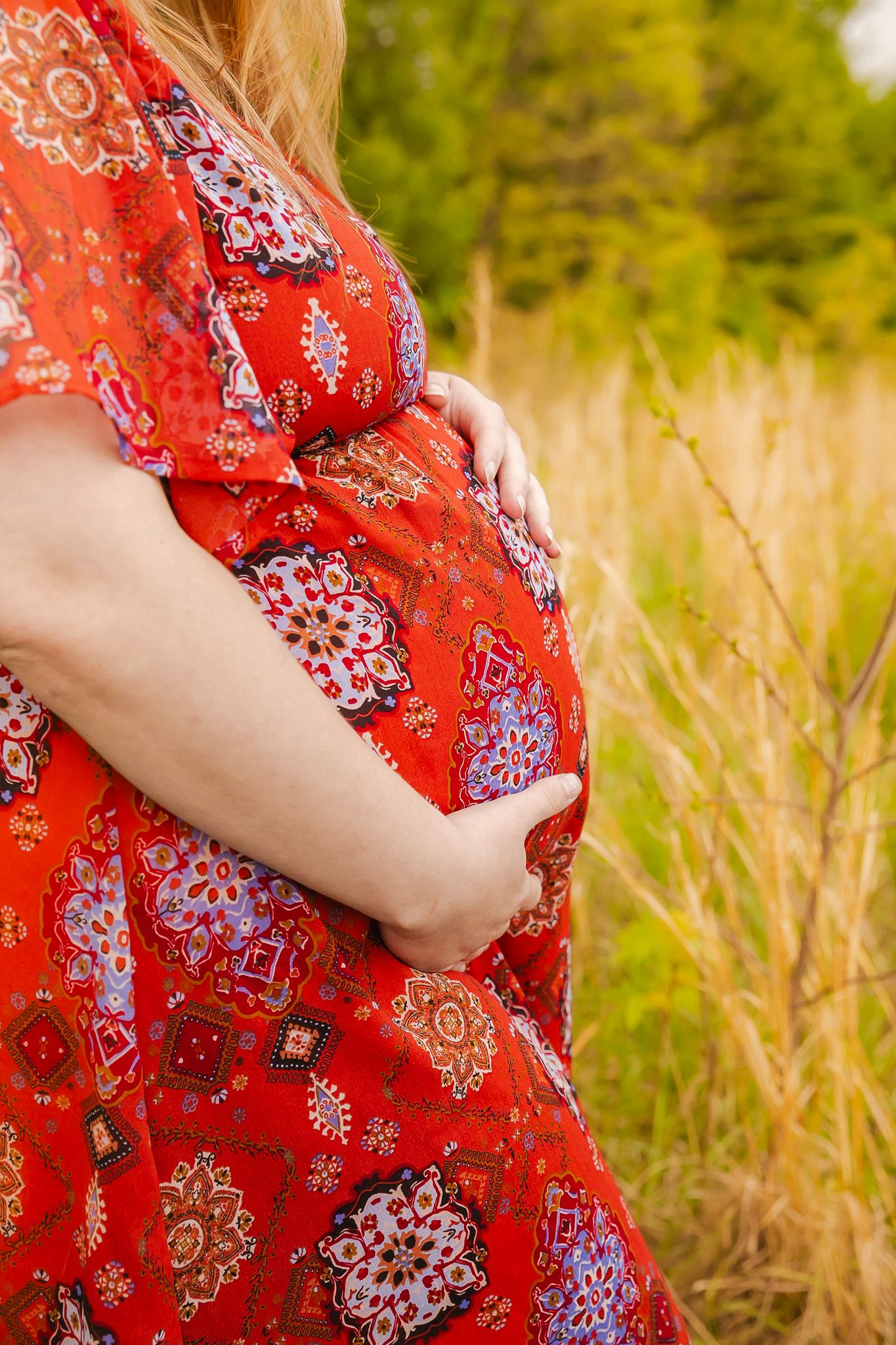 Side view of expectant mother in red floral dress holding her baby bump while standing in a grassy meadow.