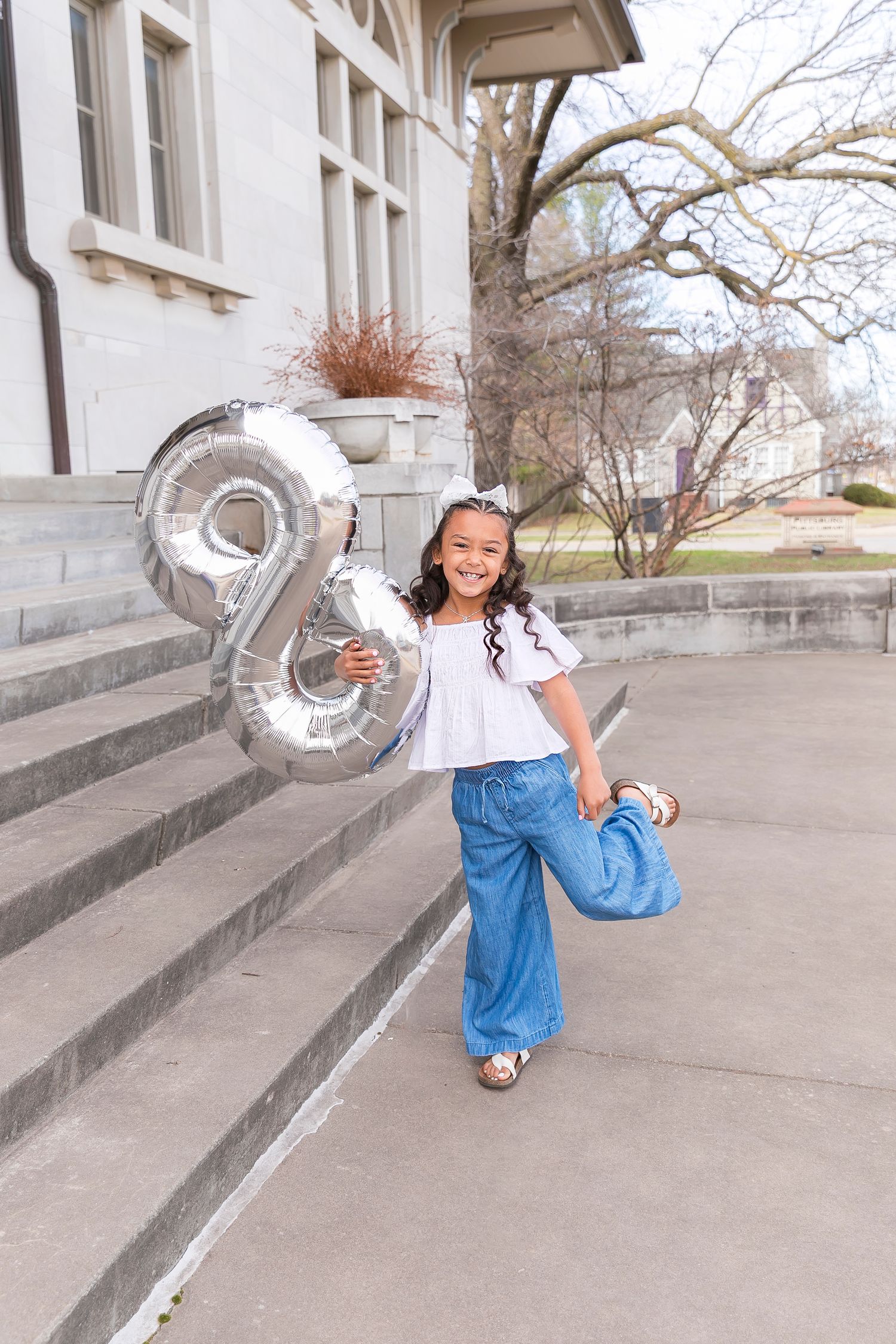 A young girl celebrates her eighth birthday standing on stone steps with silver number '8' balloons and wearing blue pants.