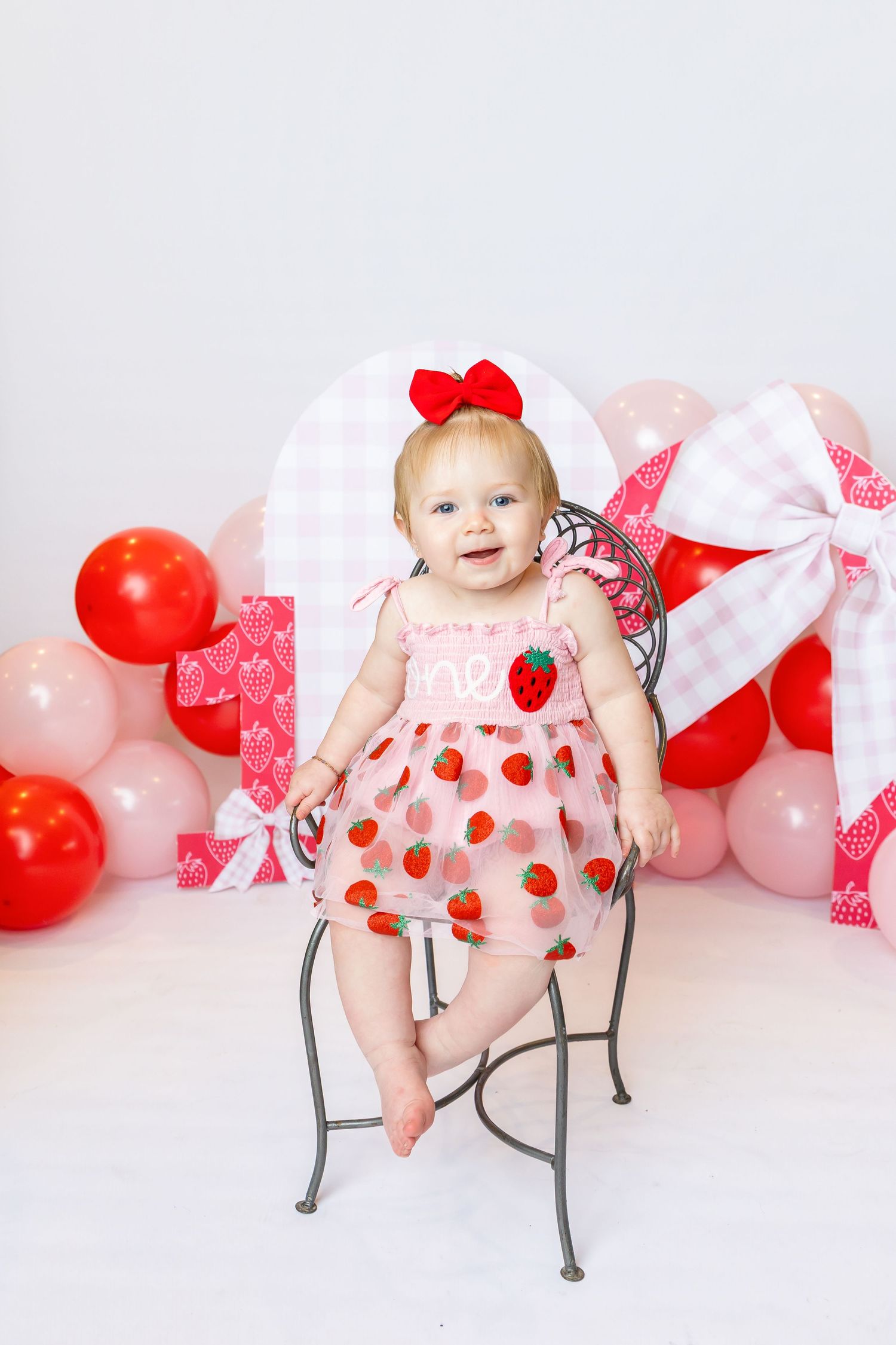 Baby sits in metal kids chair wearing a strawberry pink dress during 1st birthday shoot with family photographer Annie Robinson Photography.