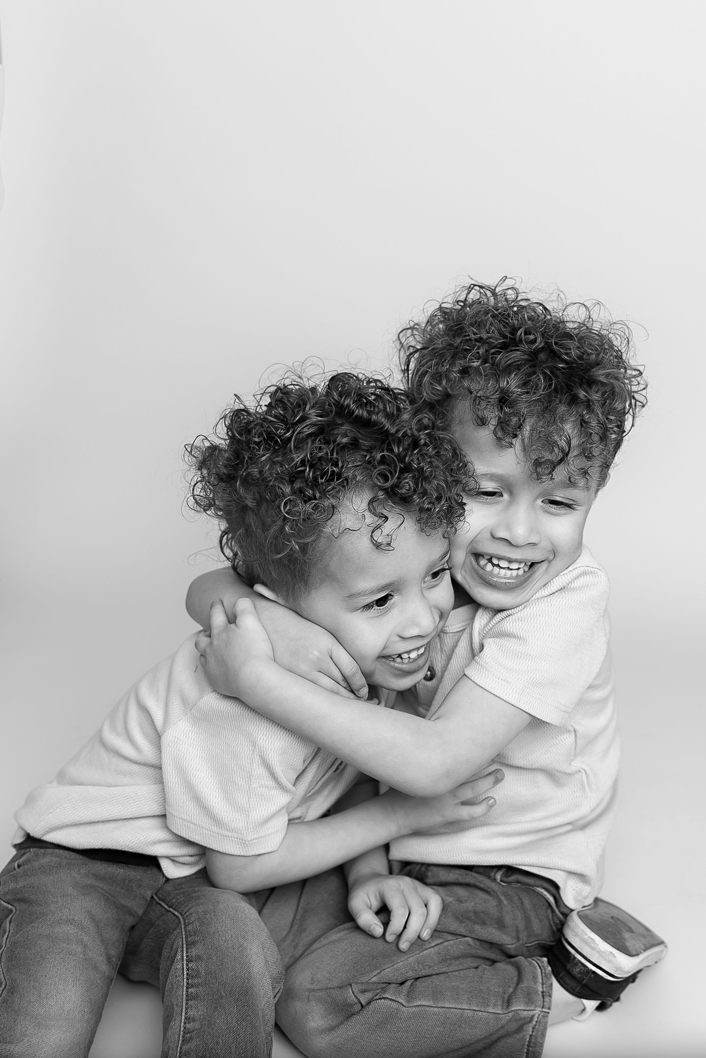 Two children, twin brothers,  with curly hair hugging and laughing joyfully in a black and white portrait.