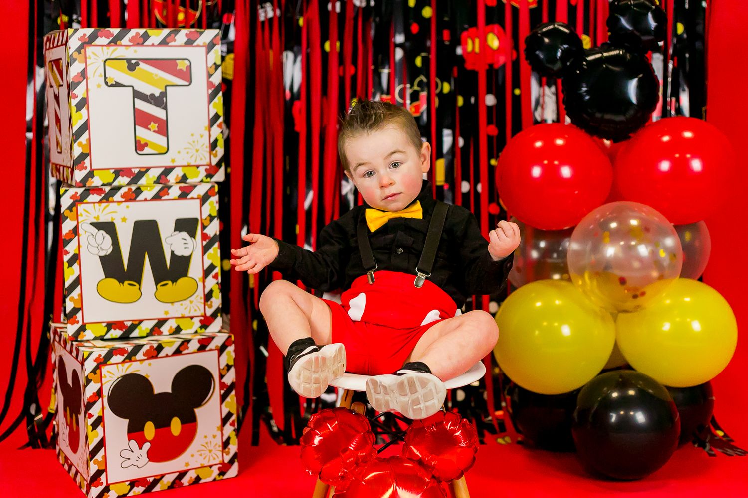 Festive Mickey Mouse themed first birthday party setup with colorful balloons and decorative blocks against a red backdrop.