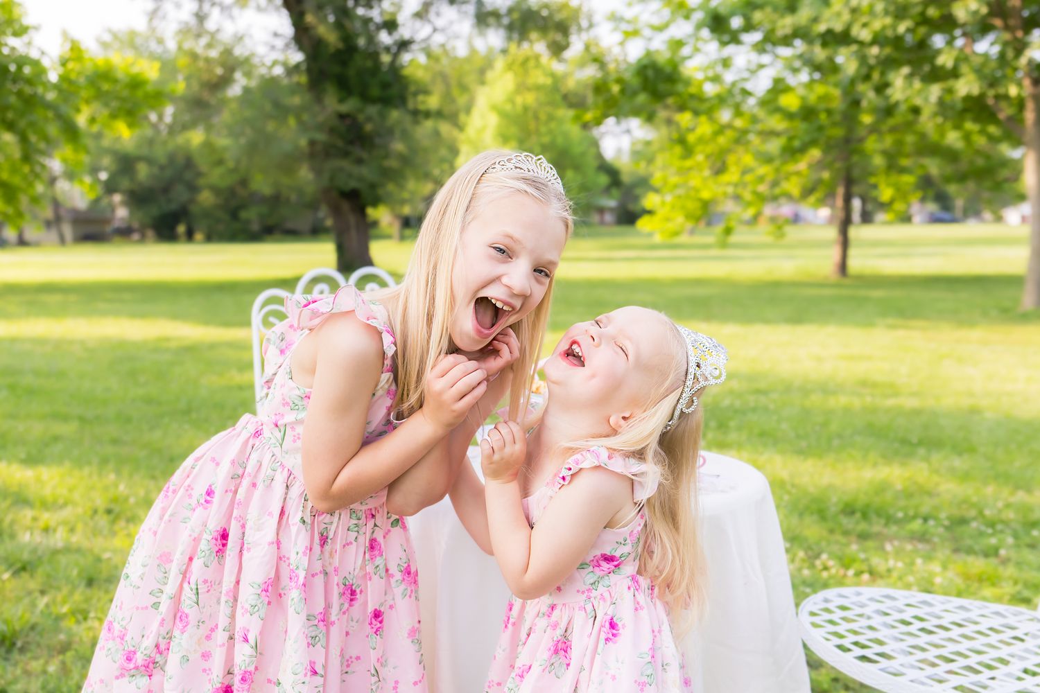 Two kids wearing matching dresses share a joyful laugh together in a sunny park during a mini session photoshoot in Pittsburg Kansas.