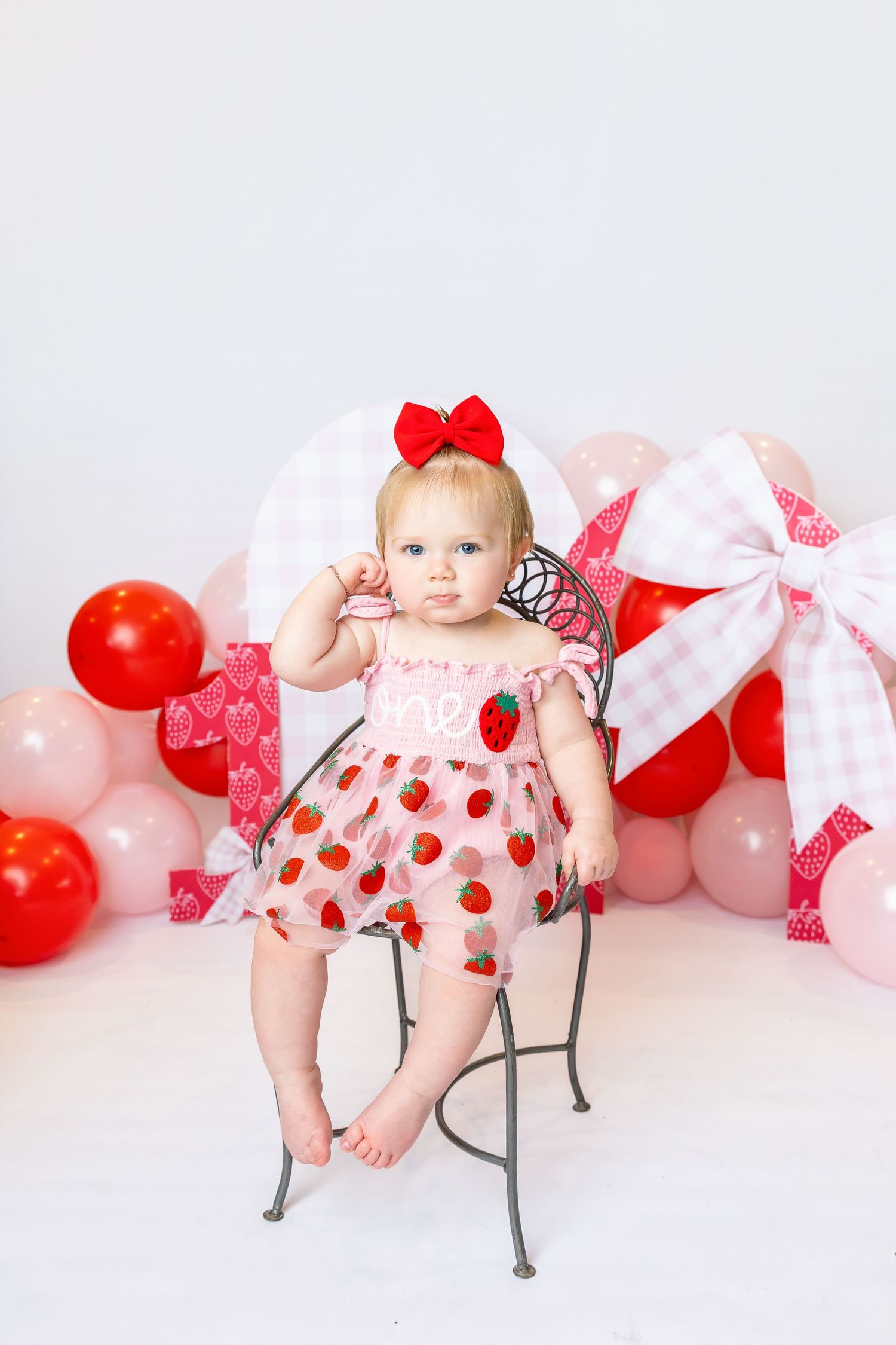 Baby sits in metal kids chair wearing a strawberry pink dress during 1st birthday shoot with family photographer Annie Robinson Photography.
