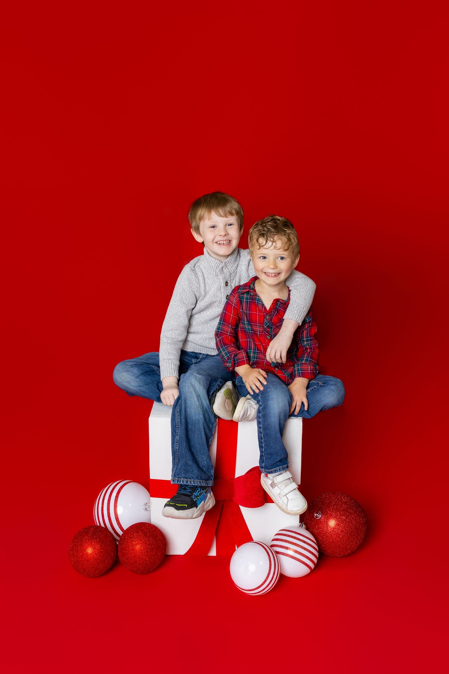 Two children in jeans sit on white present box against a bright red backdrop for a holiday portrait session in Pittsburg Ks and Joplin MO.