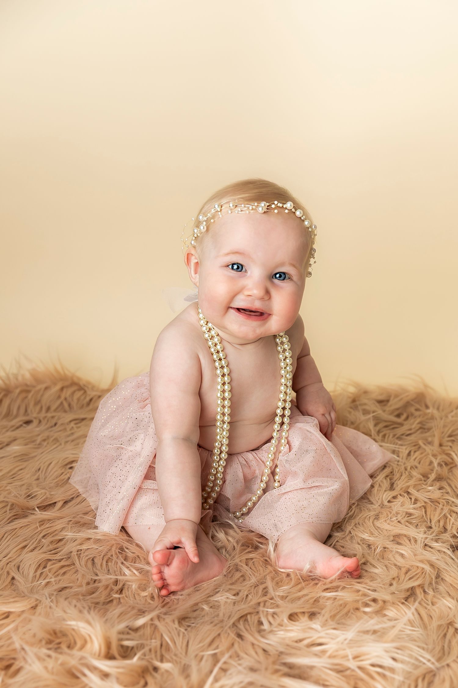 Smiling baby wearing pearls and pink tulle dress sits on fluffy beige rug during professional photo session.