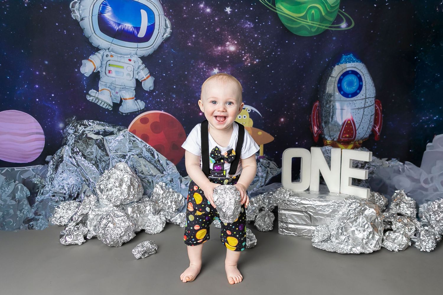 A child in a space-themed birthday photoshoot with planets, astronauts and a silver rocket against a starry backdrop.