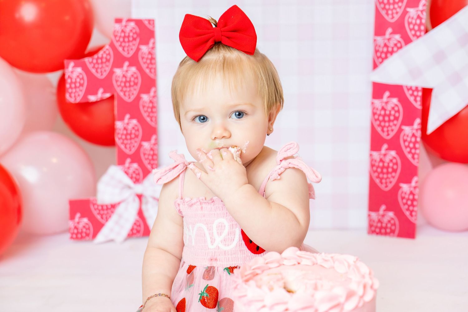 Baby wearing pink strawberry dress eats cake off her hand her during 1st birthday photoshoot with Annie Robinson Photography.