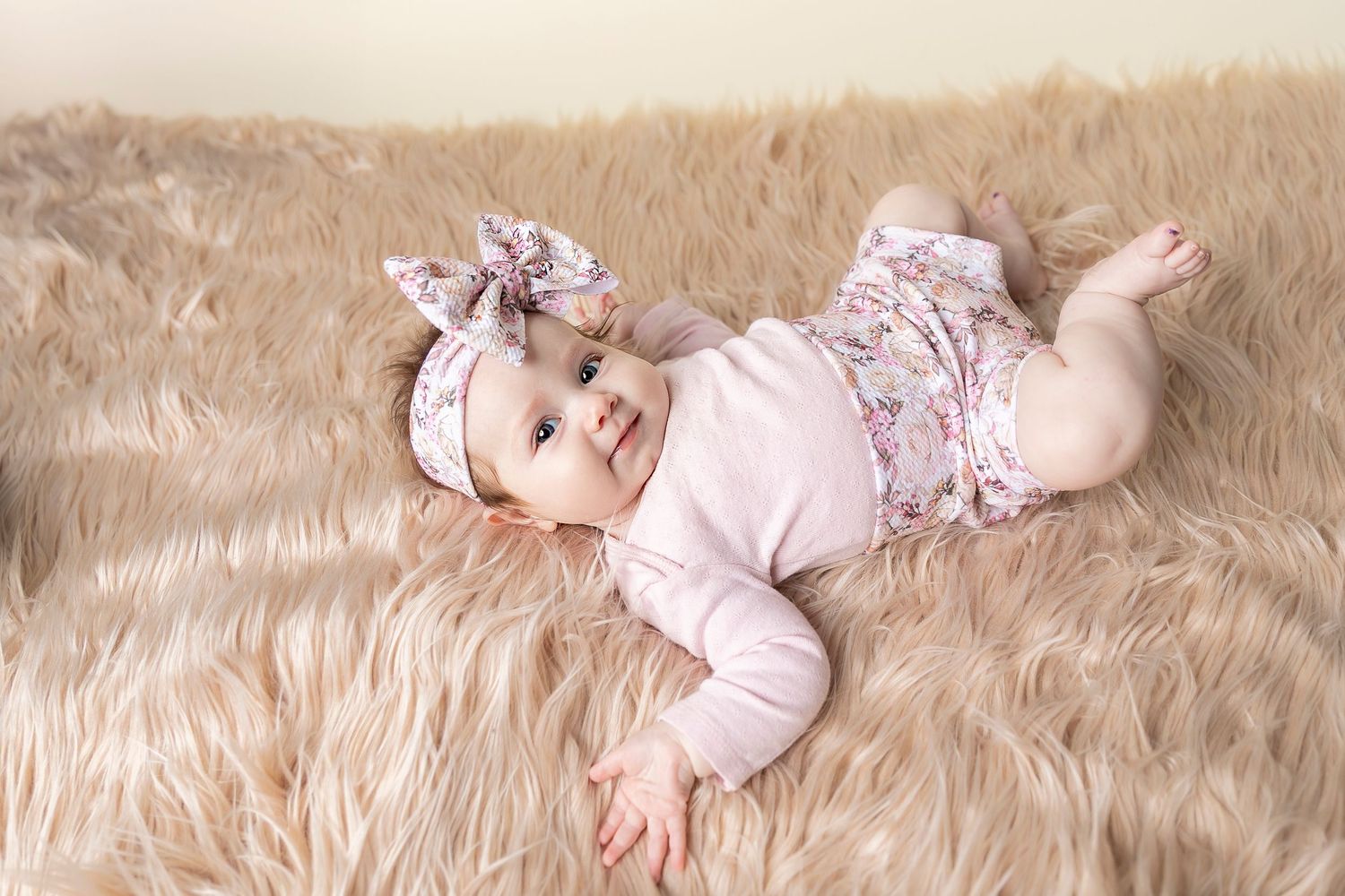 A baby wearing a pink floral outfit lies on a cream colored furry rug.