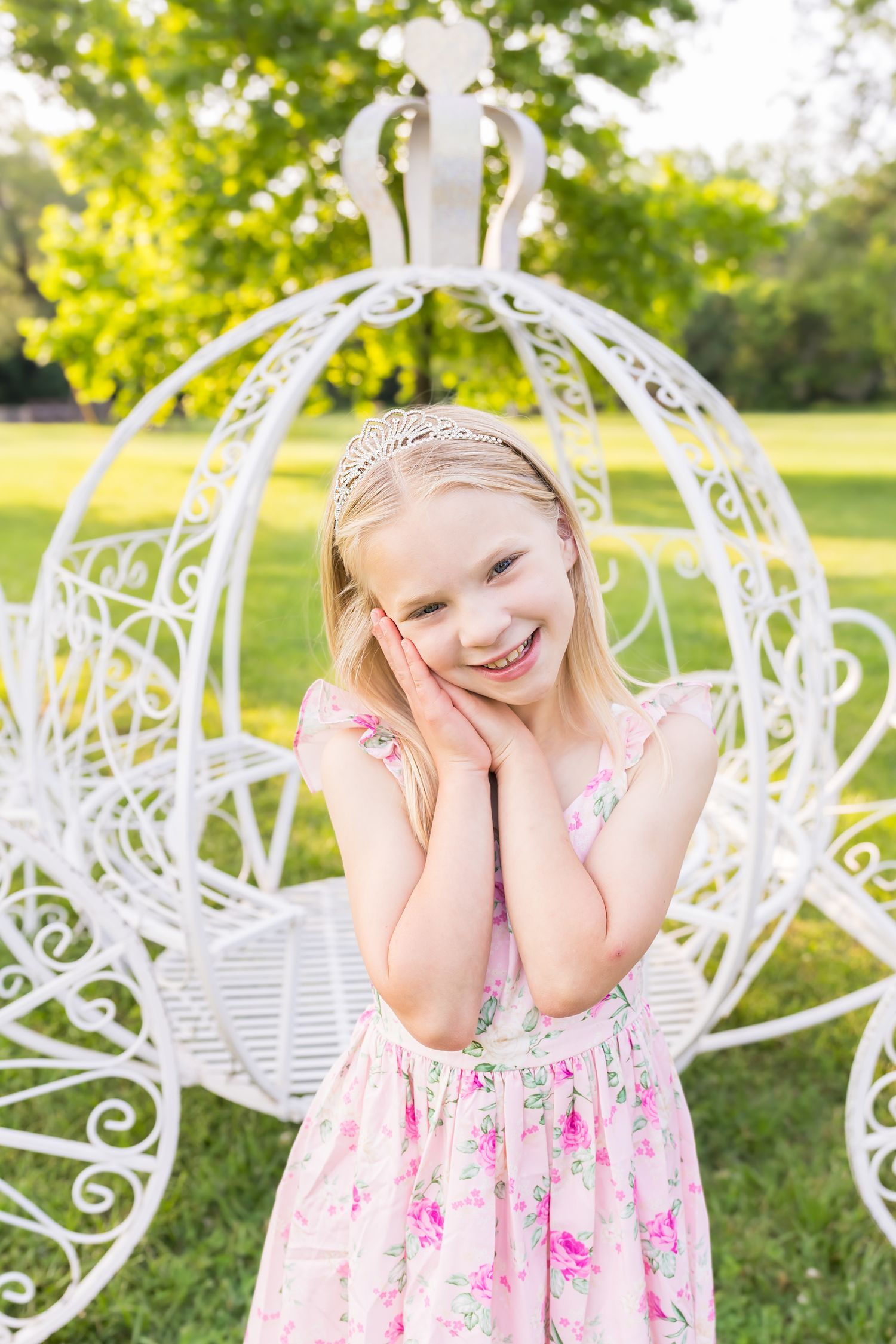 A young girl poses with hands close to face smiling in front of a large white carriage during a mini session photoshoot in Pittsburg Kansas.