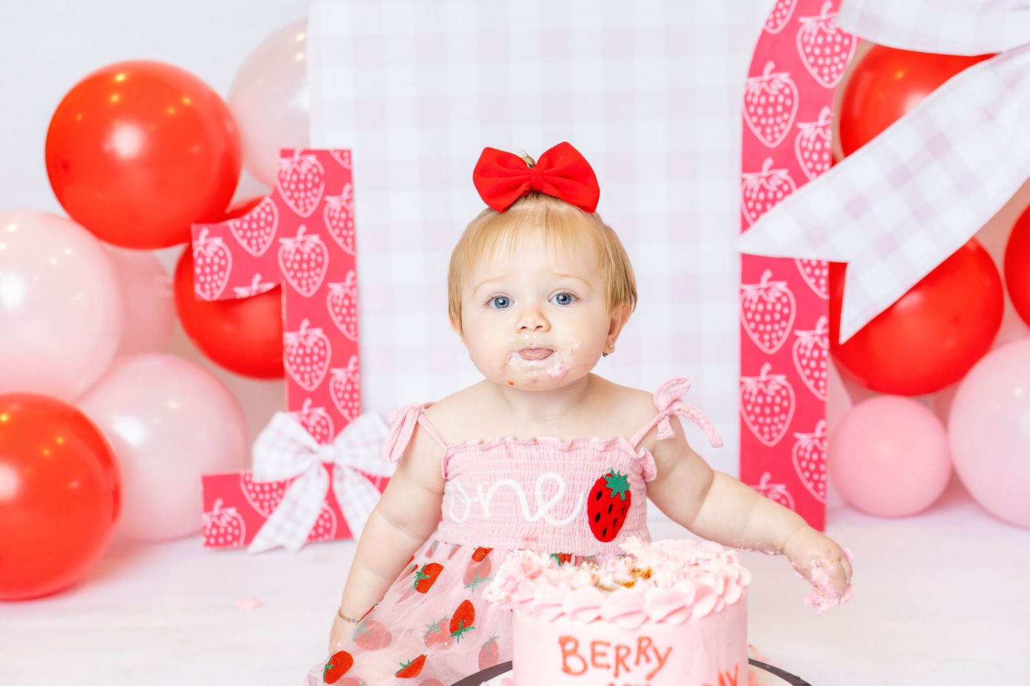 Baby wearing pink strawberry dress sits in front of cake with tongue out during 1st birthday photoshoot with Annie Robinson Photography.