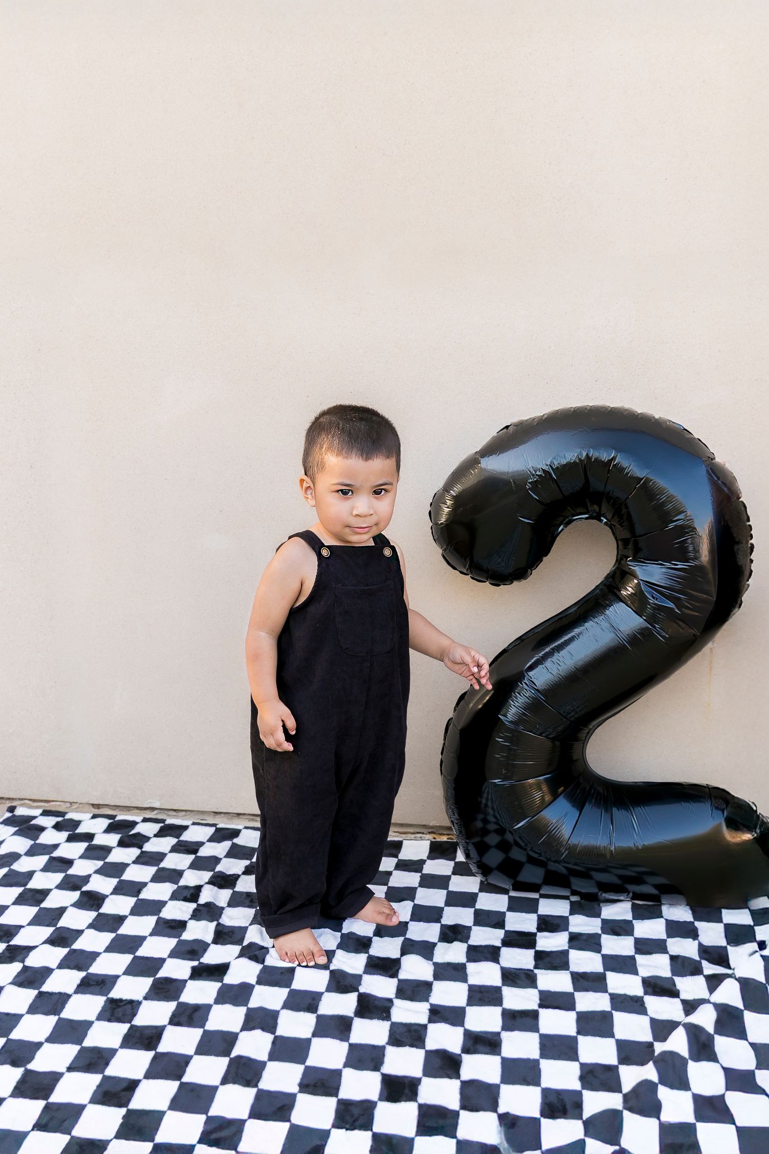 Small child in black overalls standing on checkerboard floor next to large black number 2 balloon decoration.