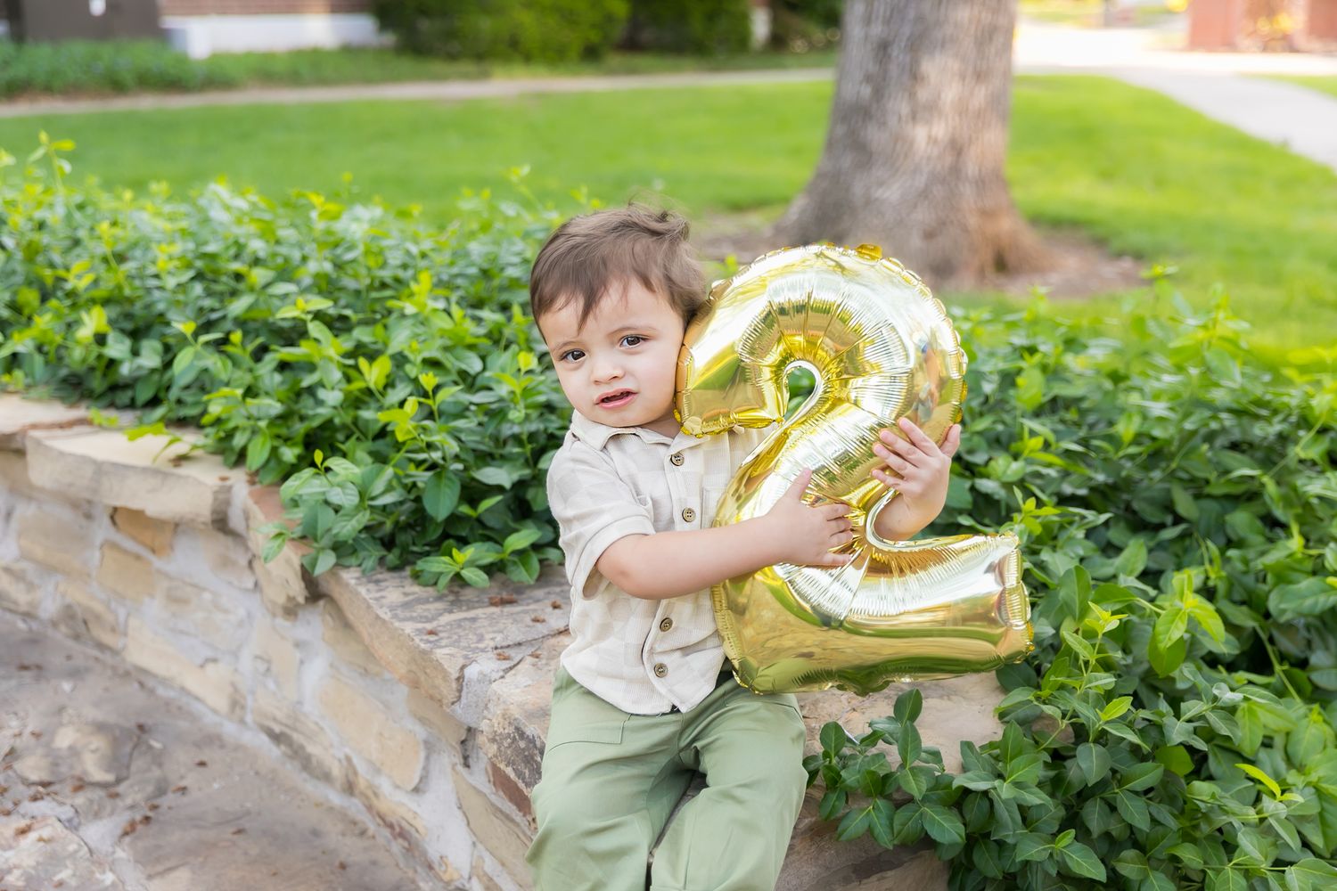 A toddler hugs a gold number 2 ballon close to his body during a 2 year old milestone photoshoot at Pitt State University in Pittsburg KS.