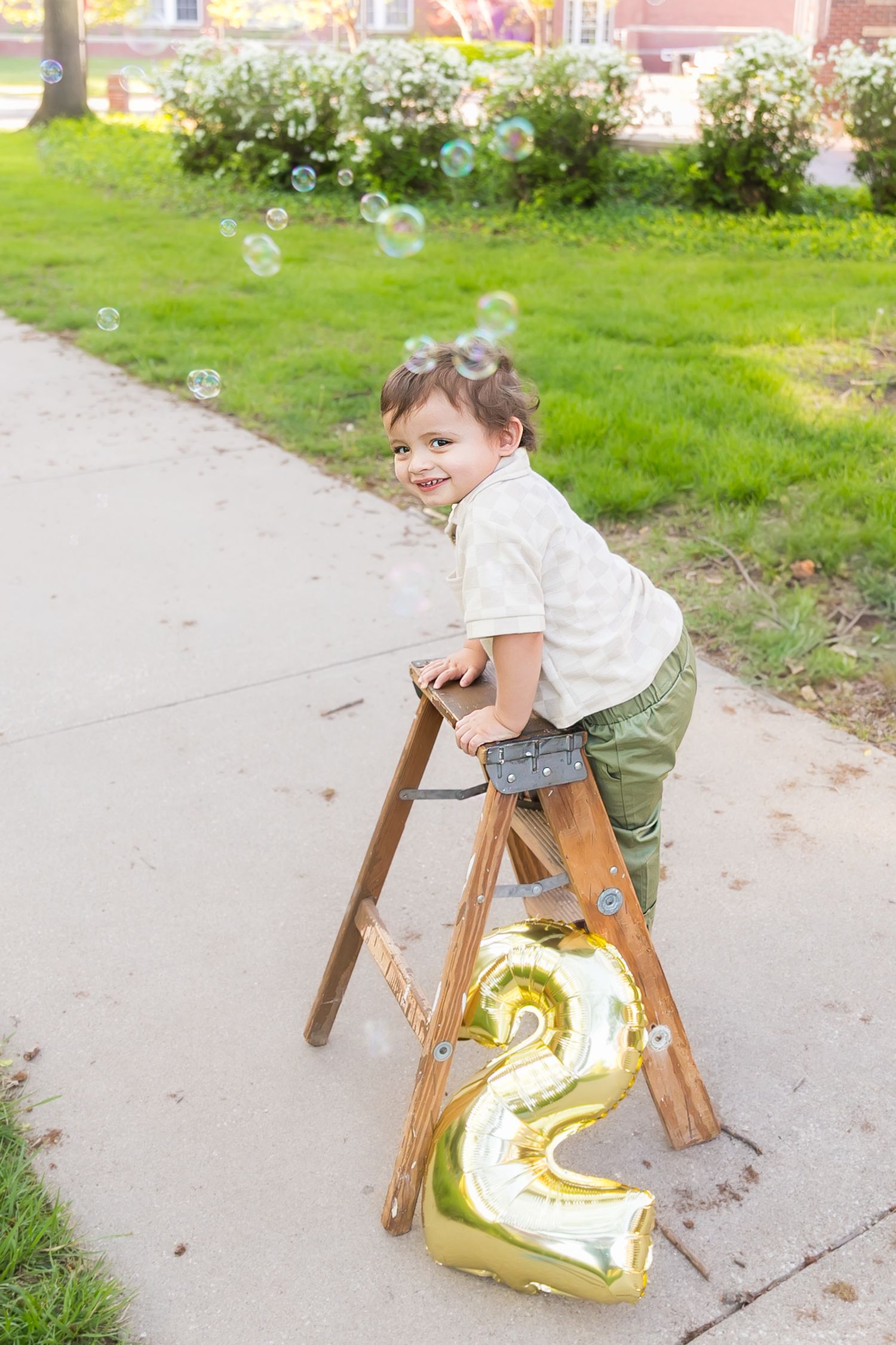 Child stands with golden number two balloon and soap bubbles decorating outdoor birthday photo scene on sidewalk at PSU in Pittsburg KS.