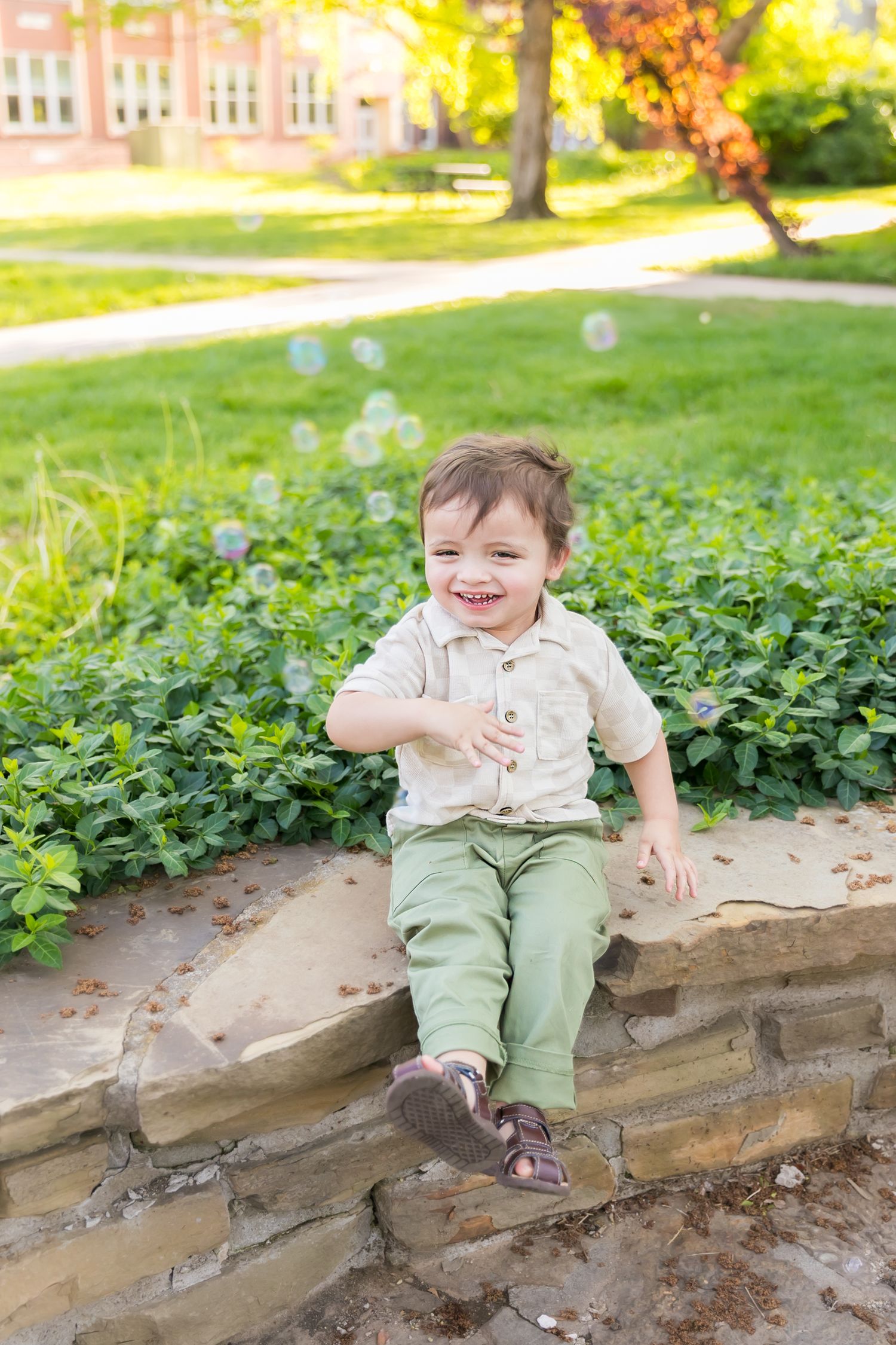 Child sits on stone wall laughing with bubbles in the air during a milestone portrait session at Pitt State University in Pittsburg KS.