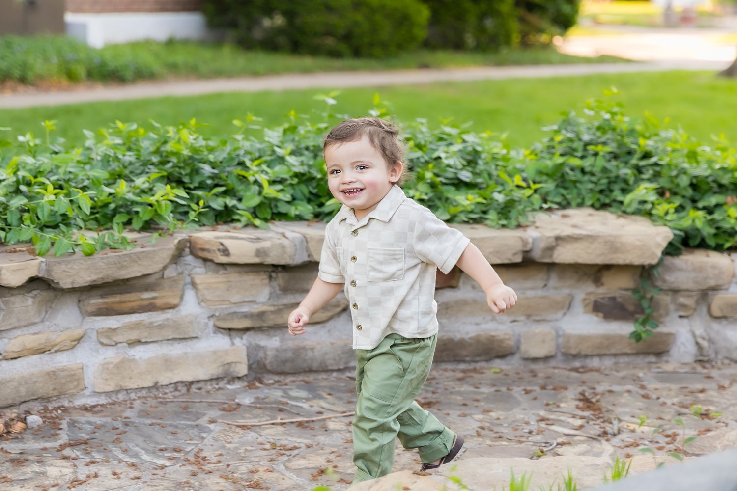 A young child plays near a stone retaining wall with green plants in the background during a milestone portrait photoshoot in Pittsburg KS.