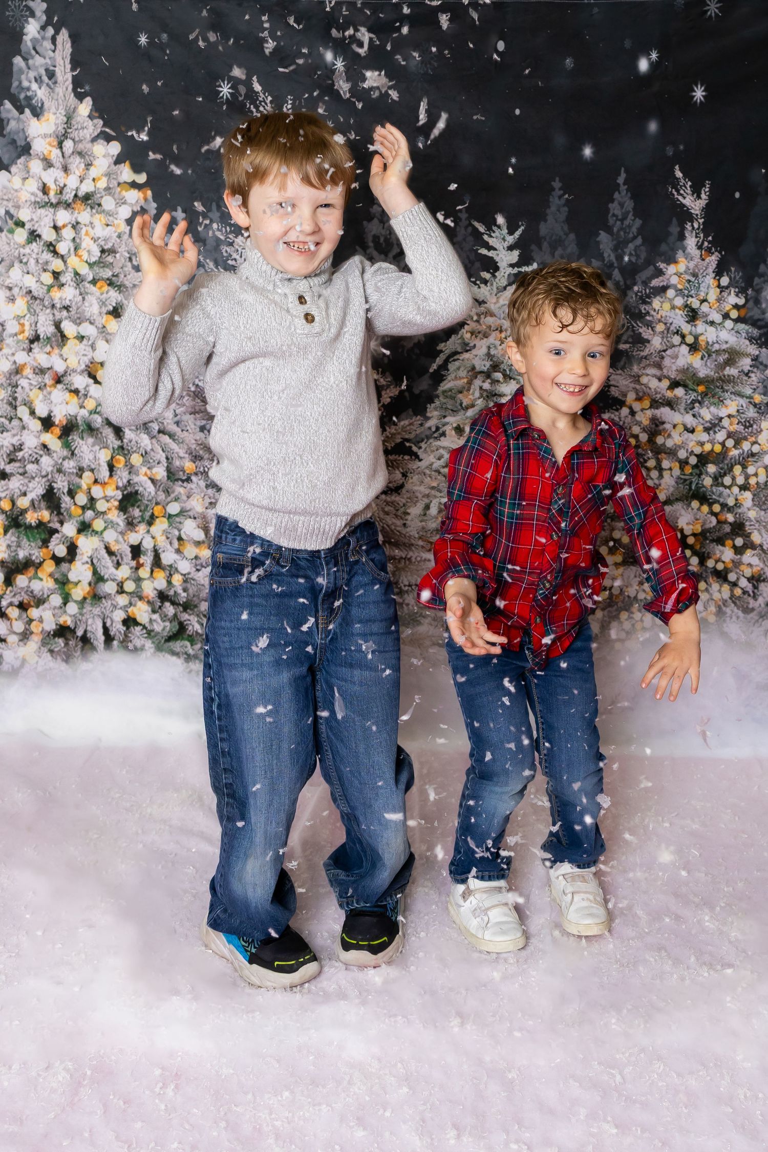 Children playing in a winter wonderland photo shoot with falling snow and decorated Christmas trees in the background.