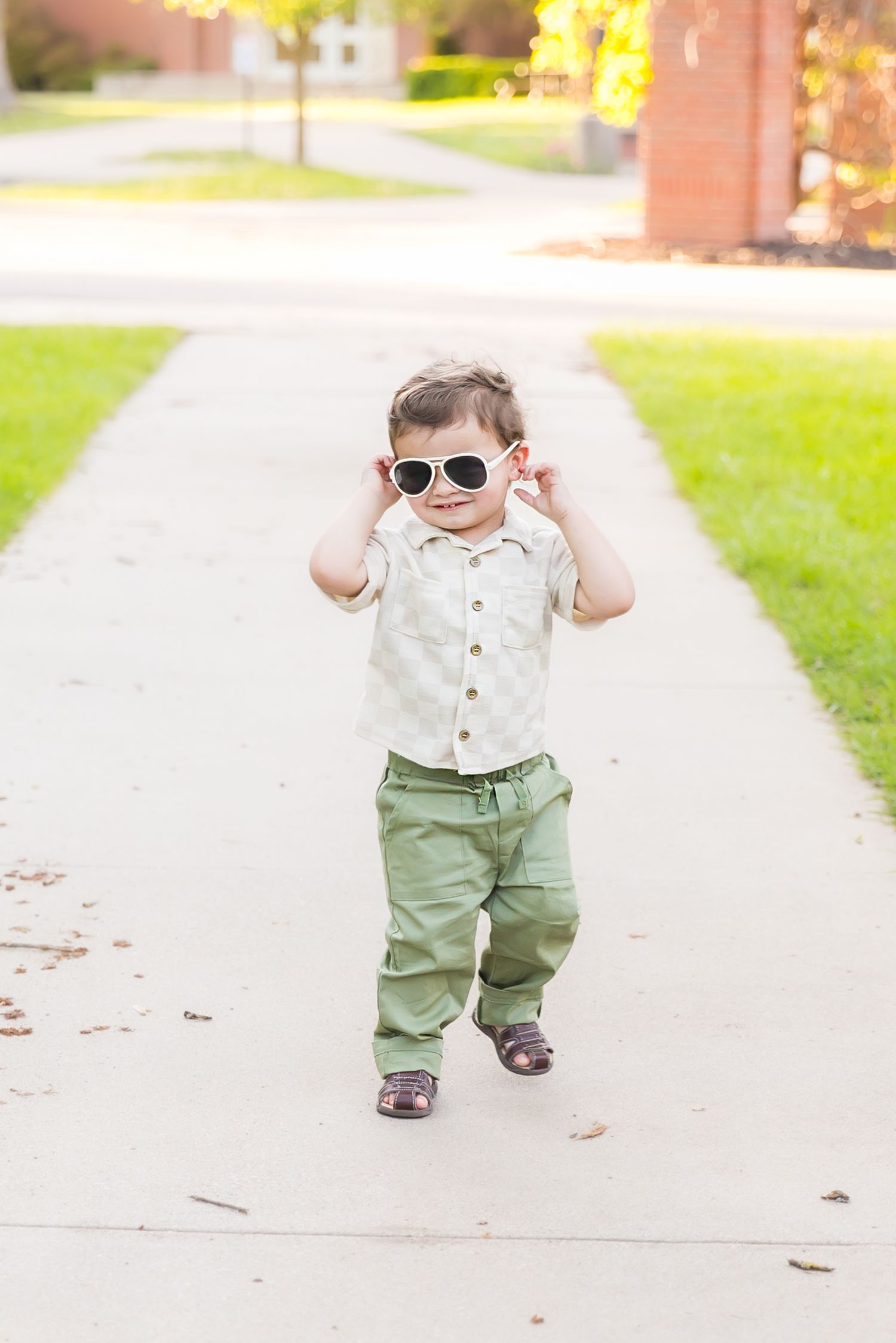 A toddler in sunglasses walks confidently down a sidewalk during a milestone birthday photoshoot at Pitt State University in Pittsburg KS.
