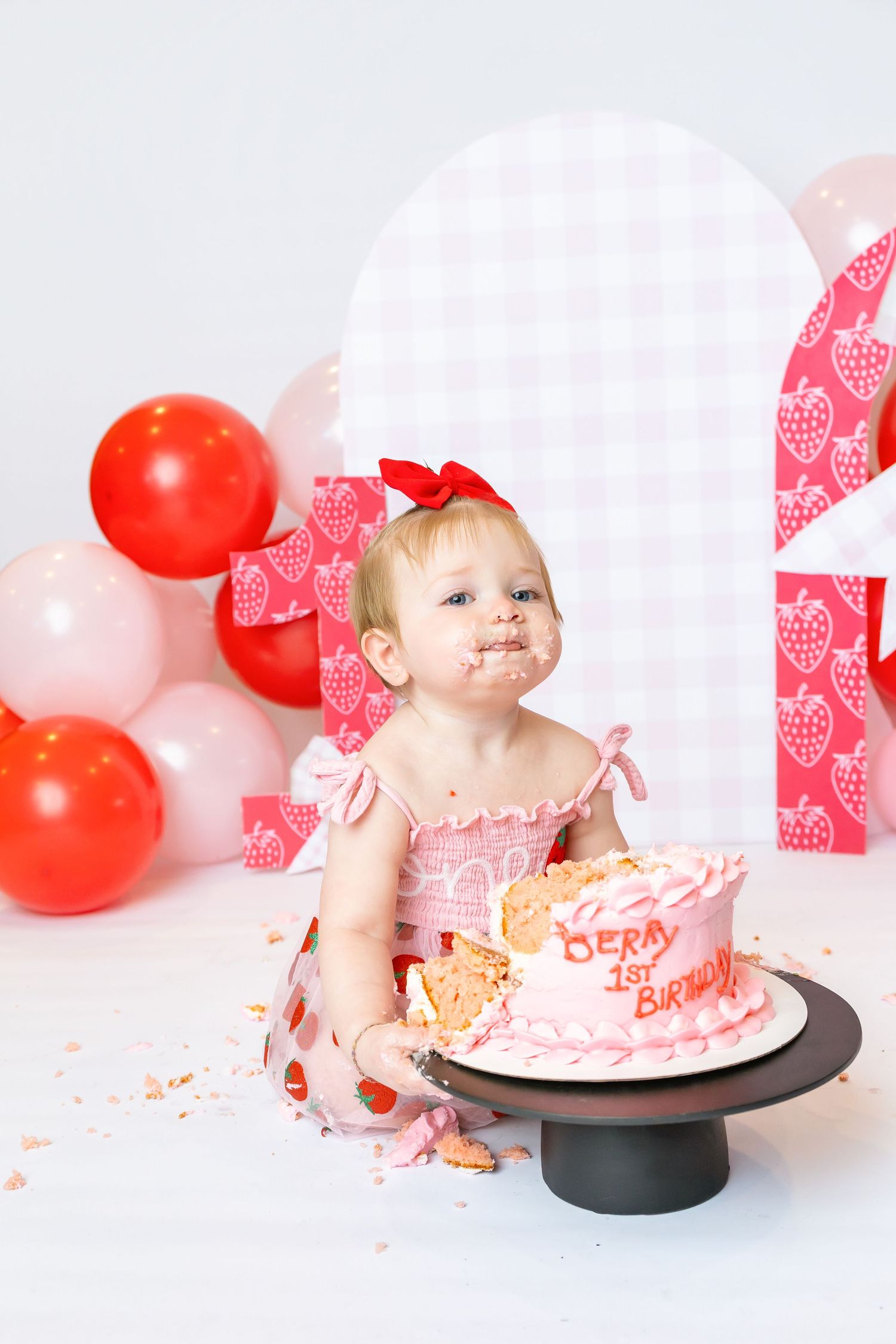 Baby wearing pink strawberry dress sits in front of smashed cake during 1st birthday photoshoot with Annie Robinson Photography.