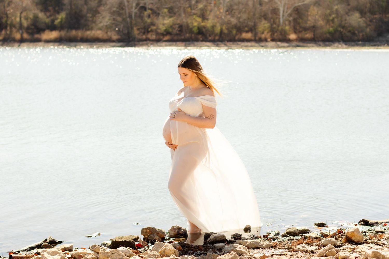 A pregnant woman stands in a lake near Austin, Texas, and holds her belly while posing for maternity photos.